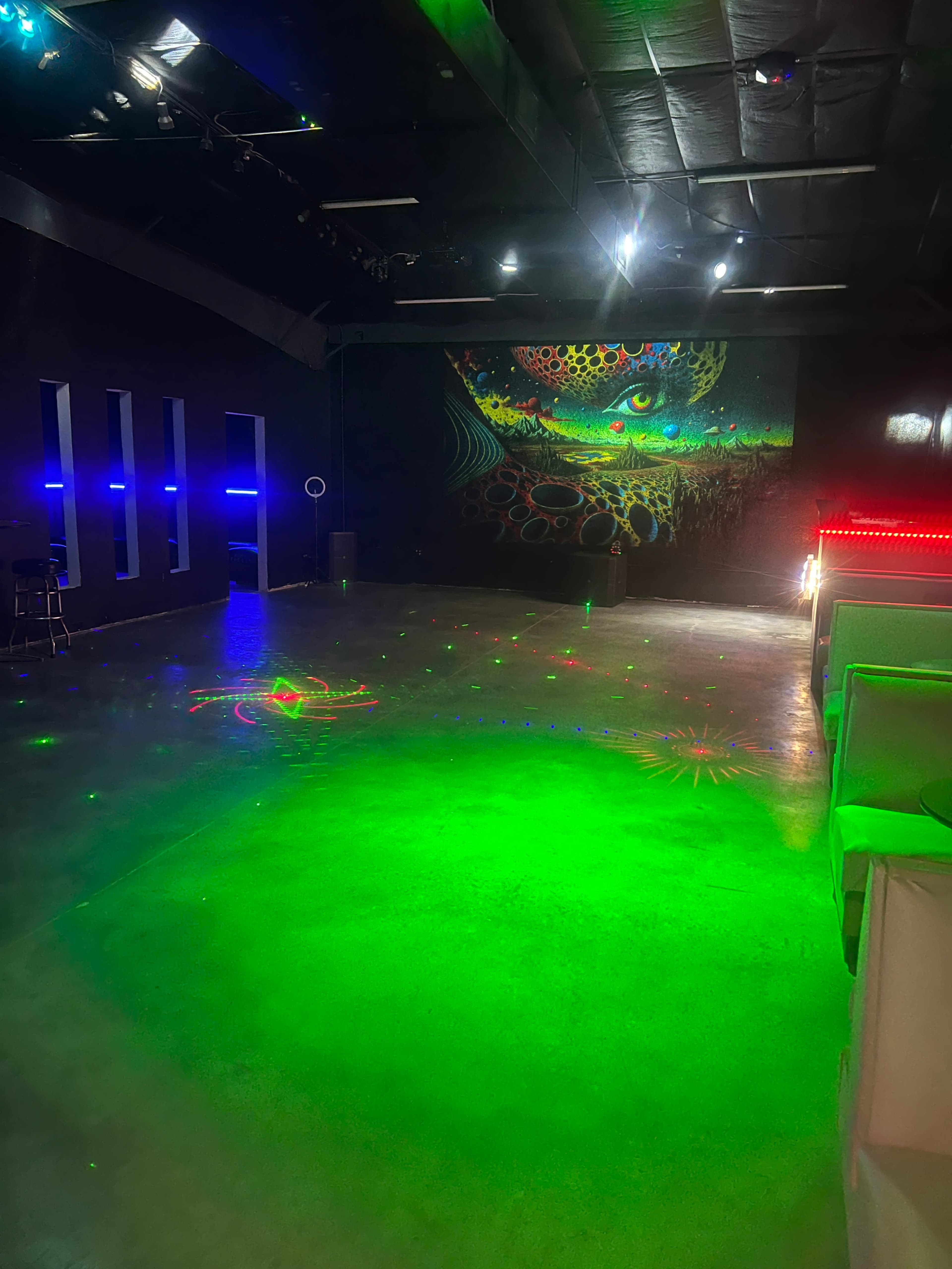 The image shows an empty indoor space with a reflective floor, colorful lighting, and a large mural on the wall.