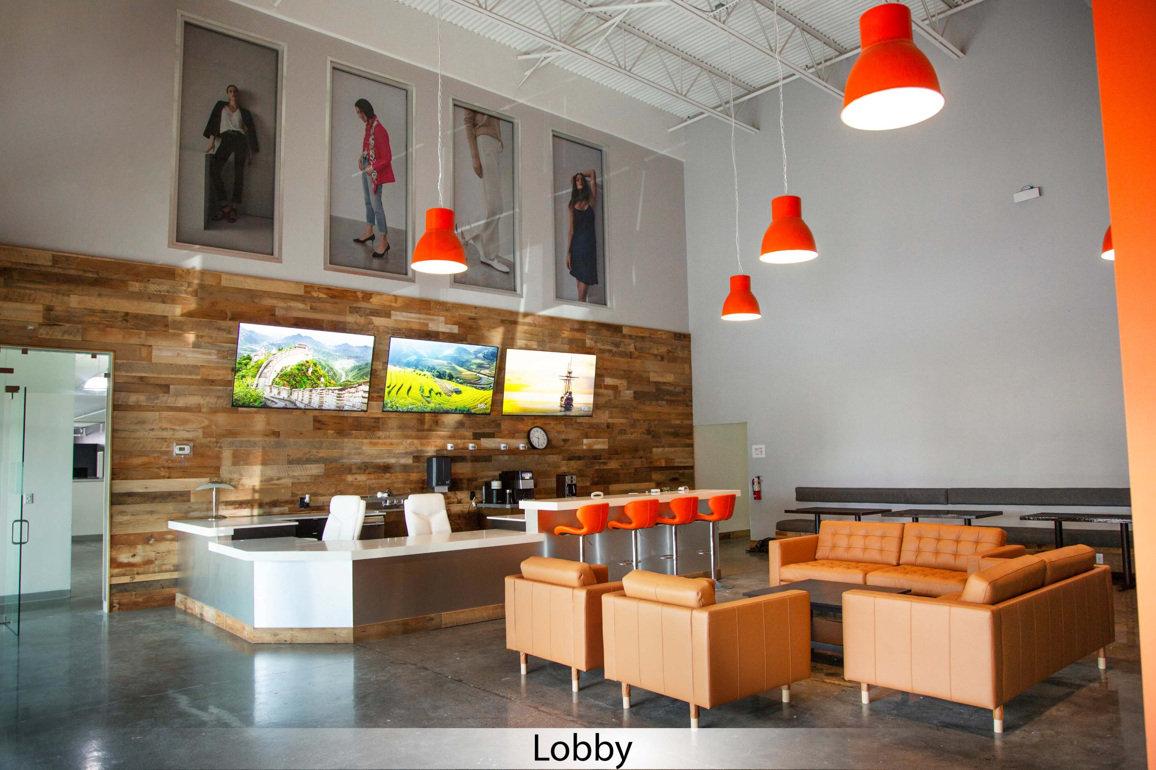 The image shows a modern lobby featuring a sleek reception area, orange pendant lights, a brown wooden accent wall, and a seating arrangement with orange sofas and chairs.