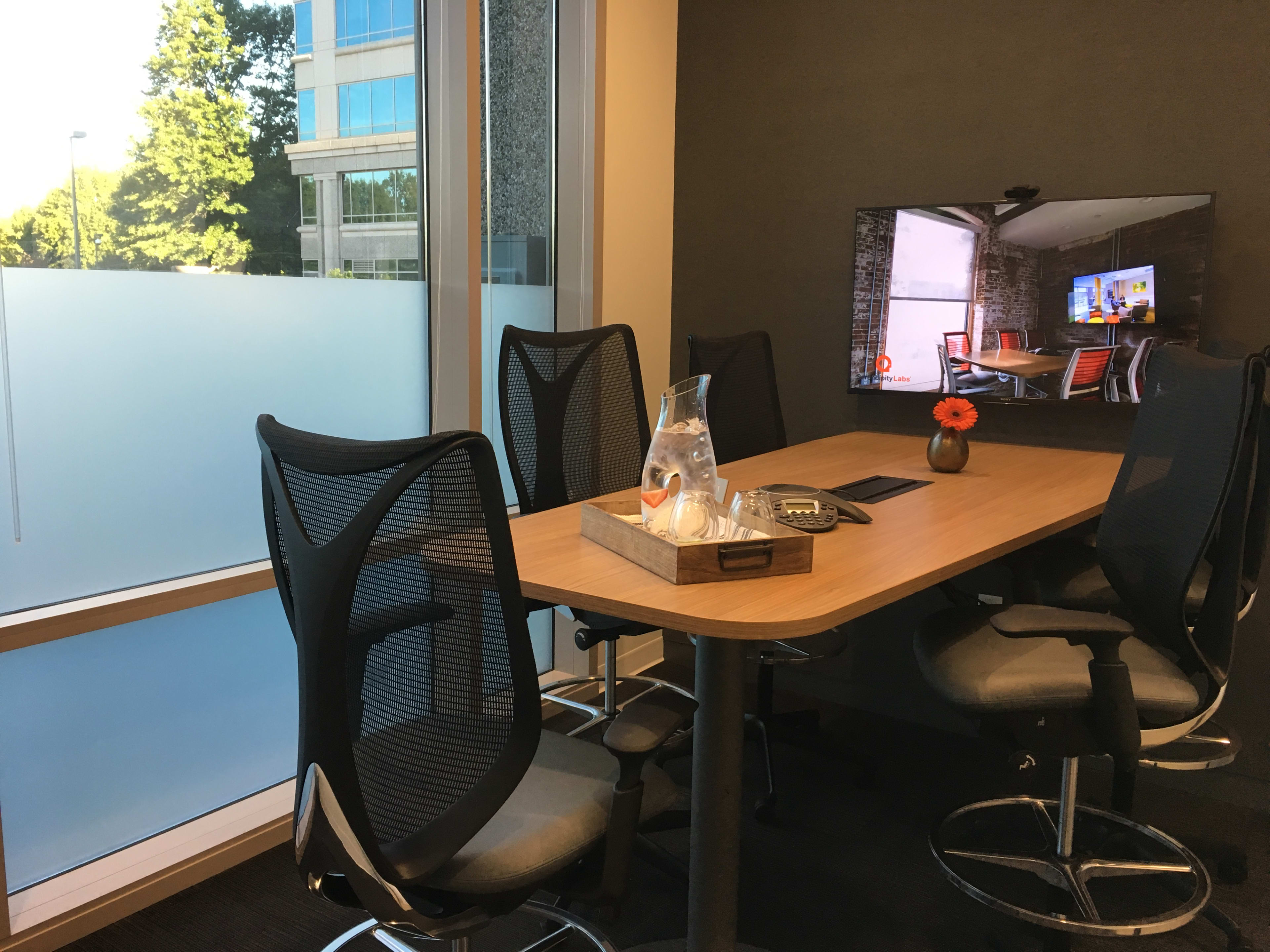 A modern conference room features a wooden table surrounded by ergonomic chairs and a wall-mounted television displaying a presentation.