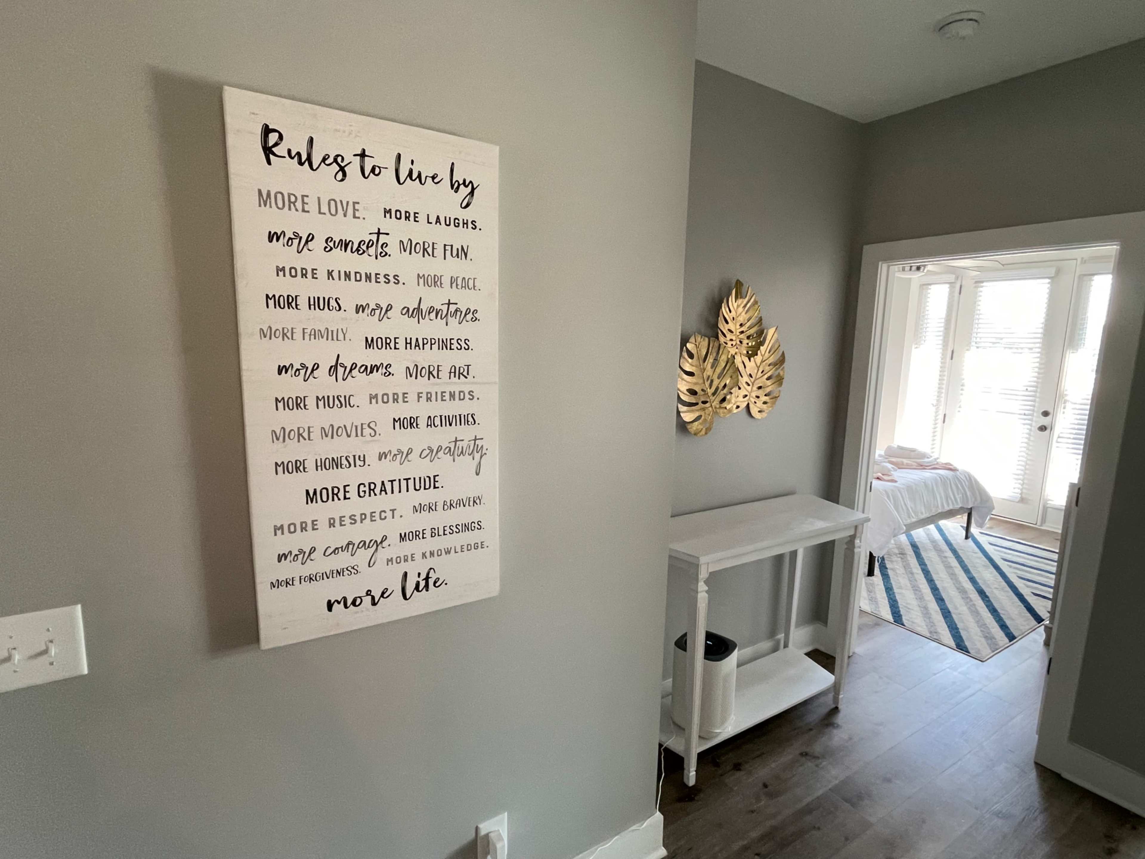 A hallway features a framed list of "Rules to Live By" on the wall, with a console table and decorative leaves nearby, leading to a well-lit room.