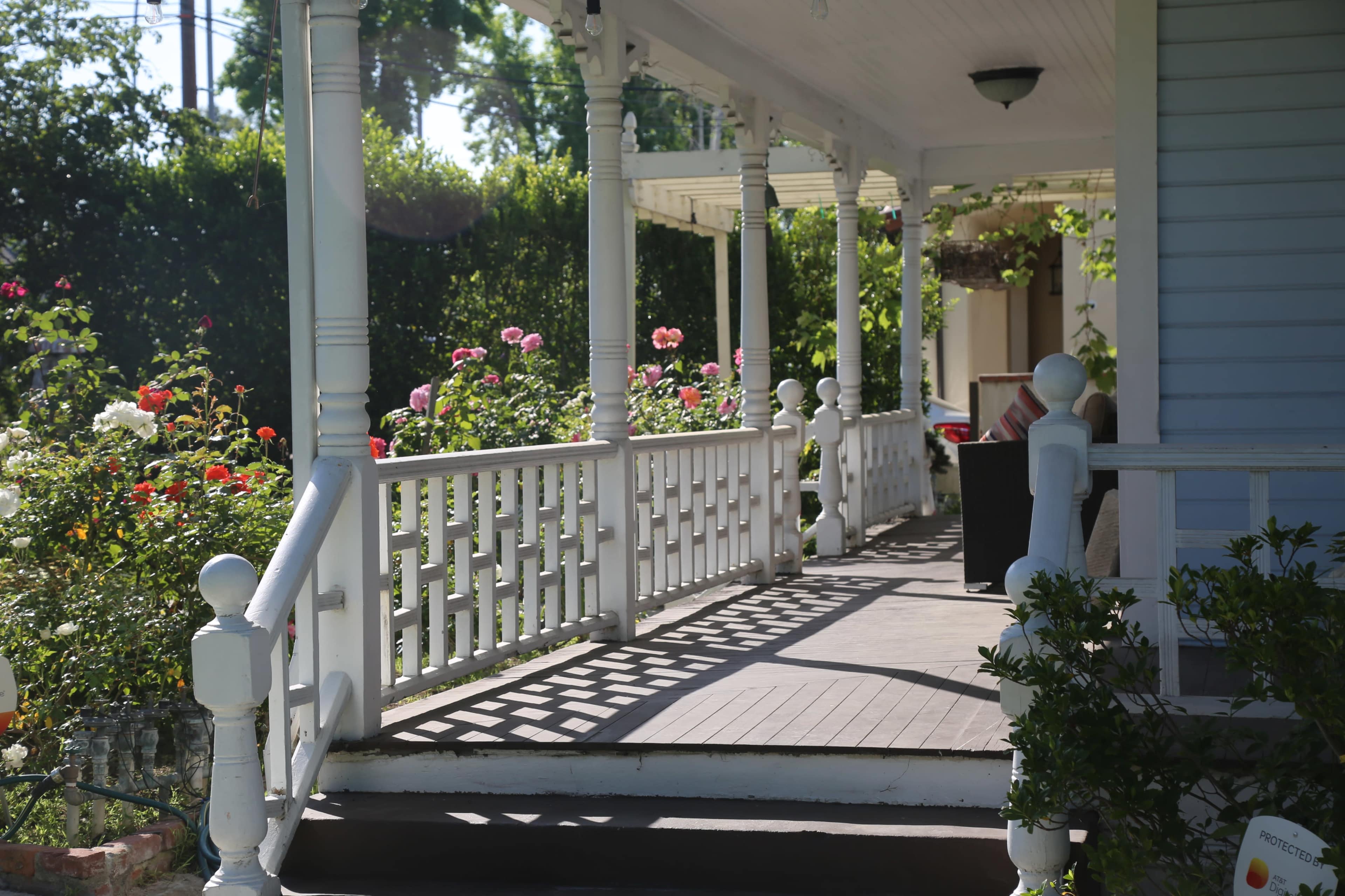 A covered porch with white railings and a wooden floor, surrounded by blooming rose bushes.
