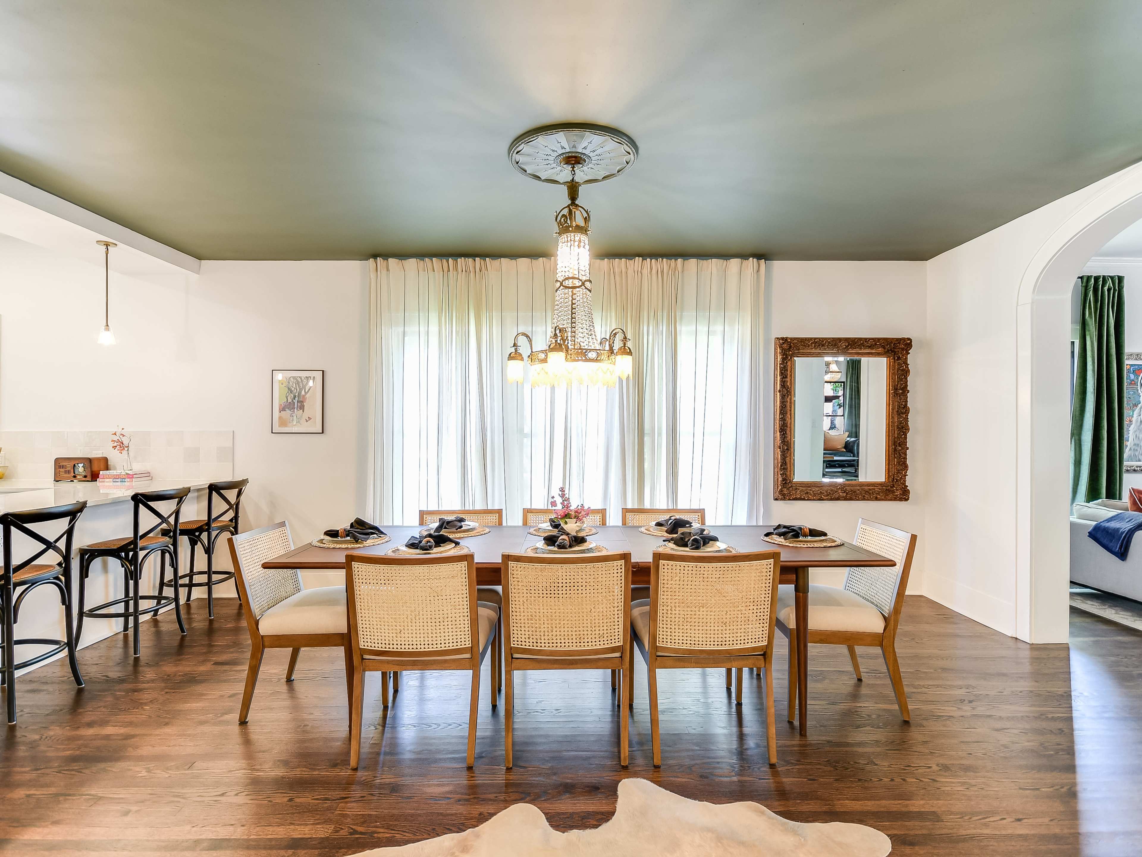 A dining area features a large wooden table surrounded by six chairs, with a chandelier above and large windows draped with light curtains.