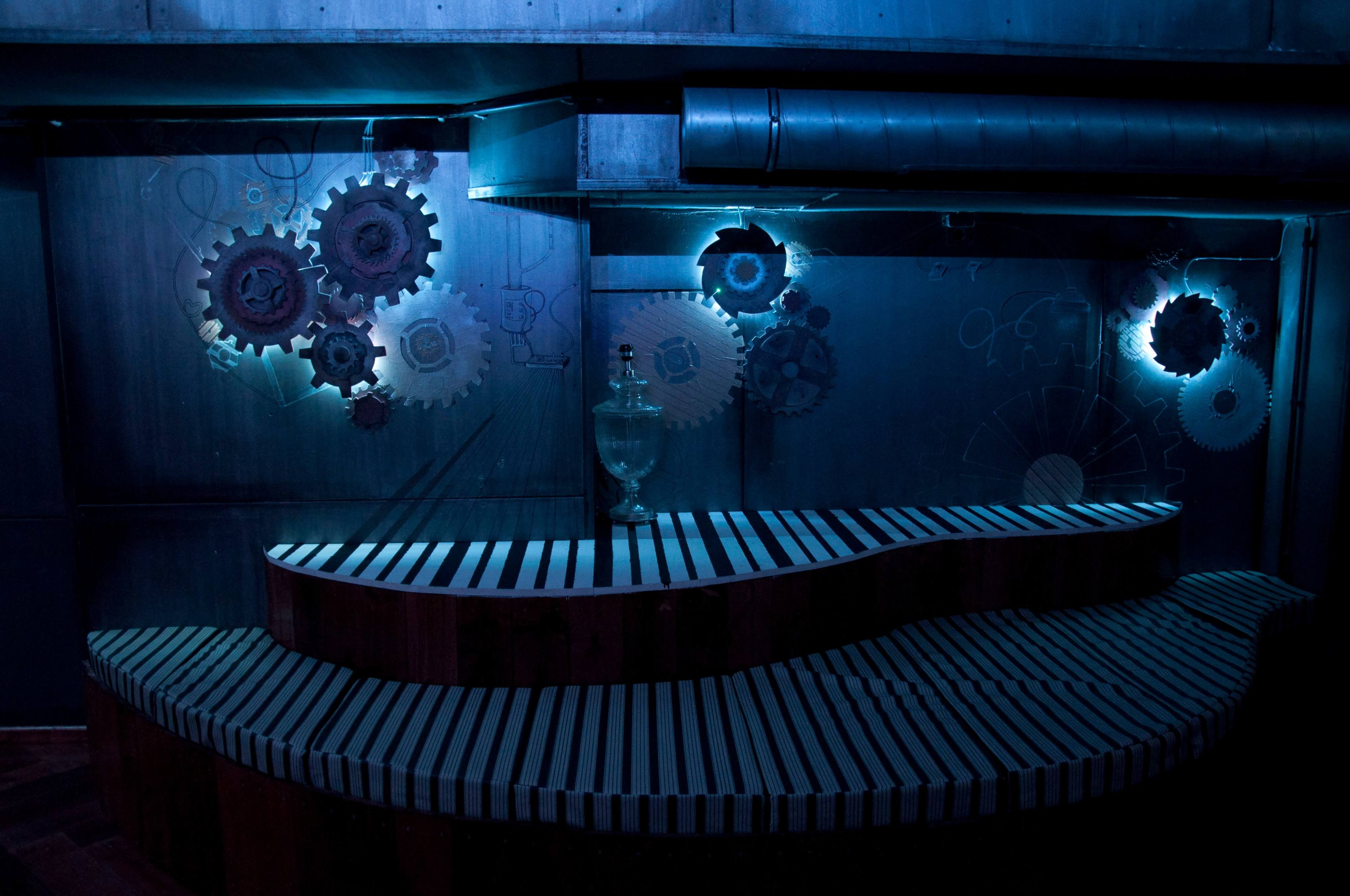 The image shows a dimly lit bar area with a curved wooden counter and illuminated wall decorations featuring metallic gears.