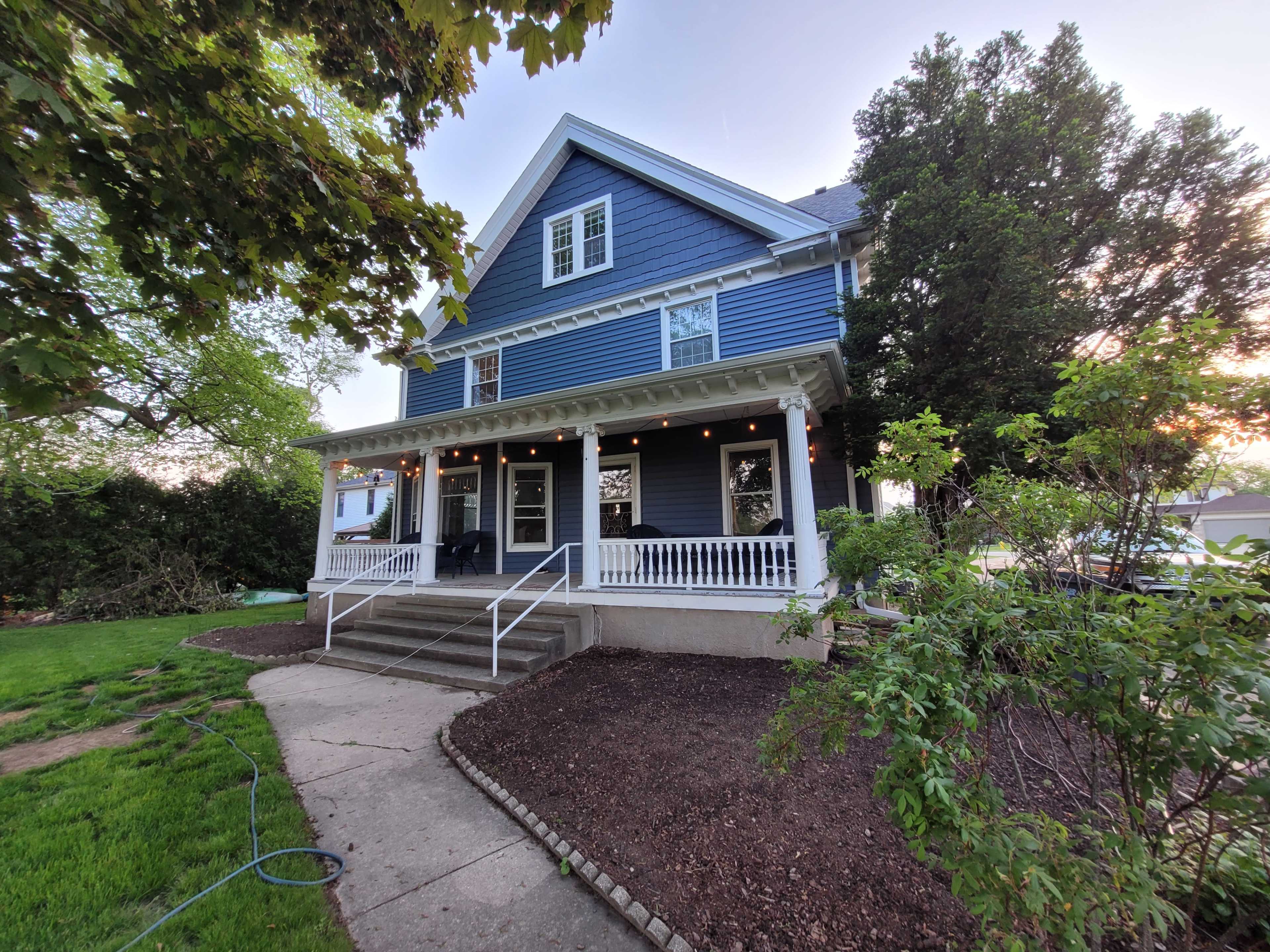 A three-story blue house with a front porch is surrounded by greenery and features outdoor lighting.