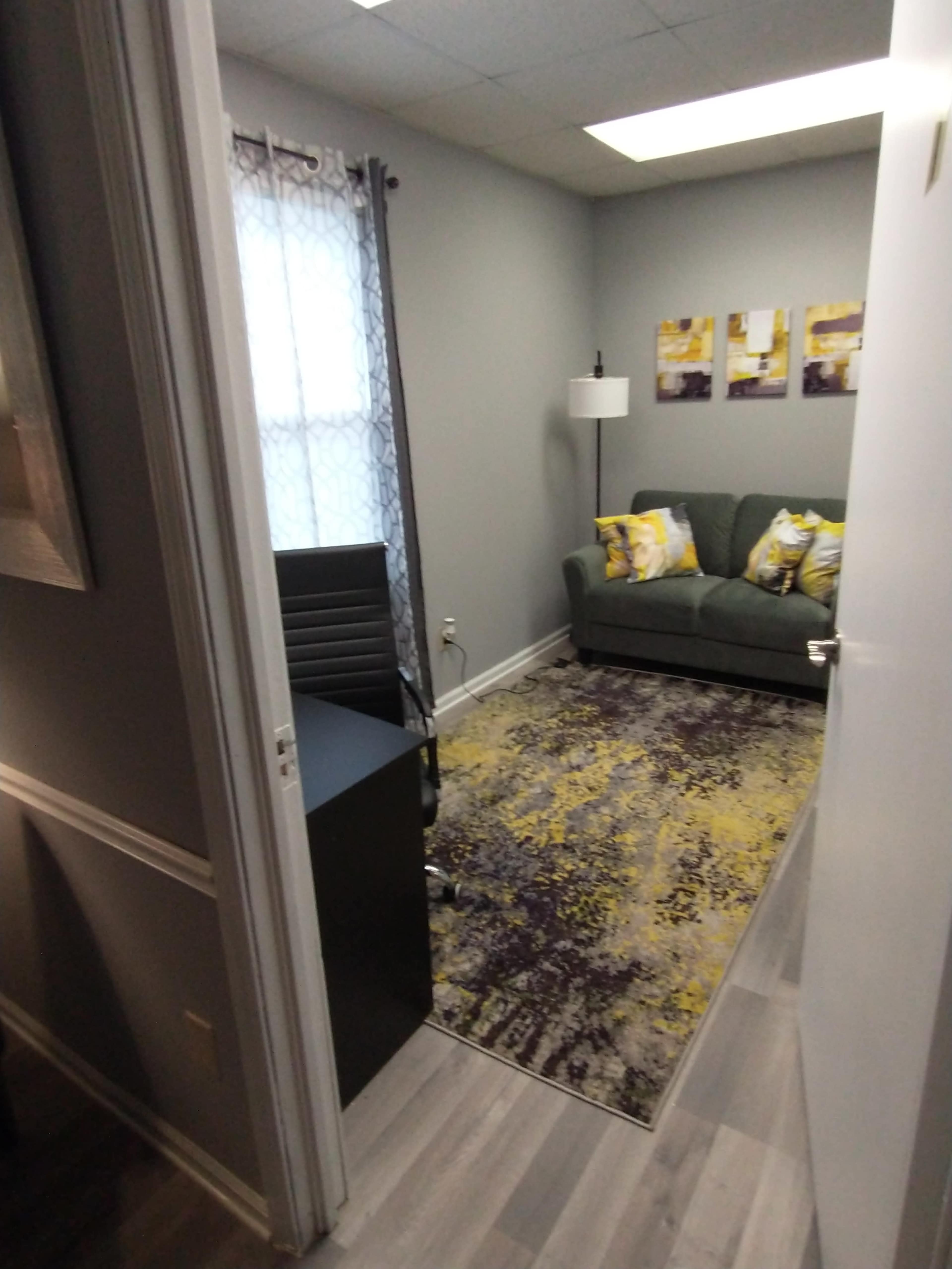 An office space featuring a small sitting area with a couch, a lamp, and a patterned rug, along with a desk near the entrance.