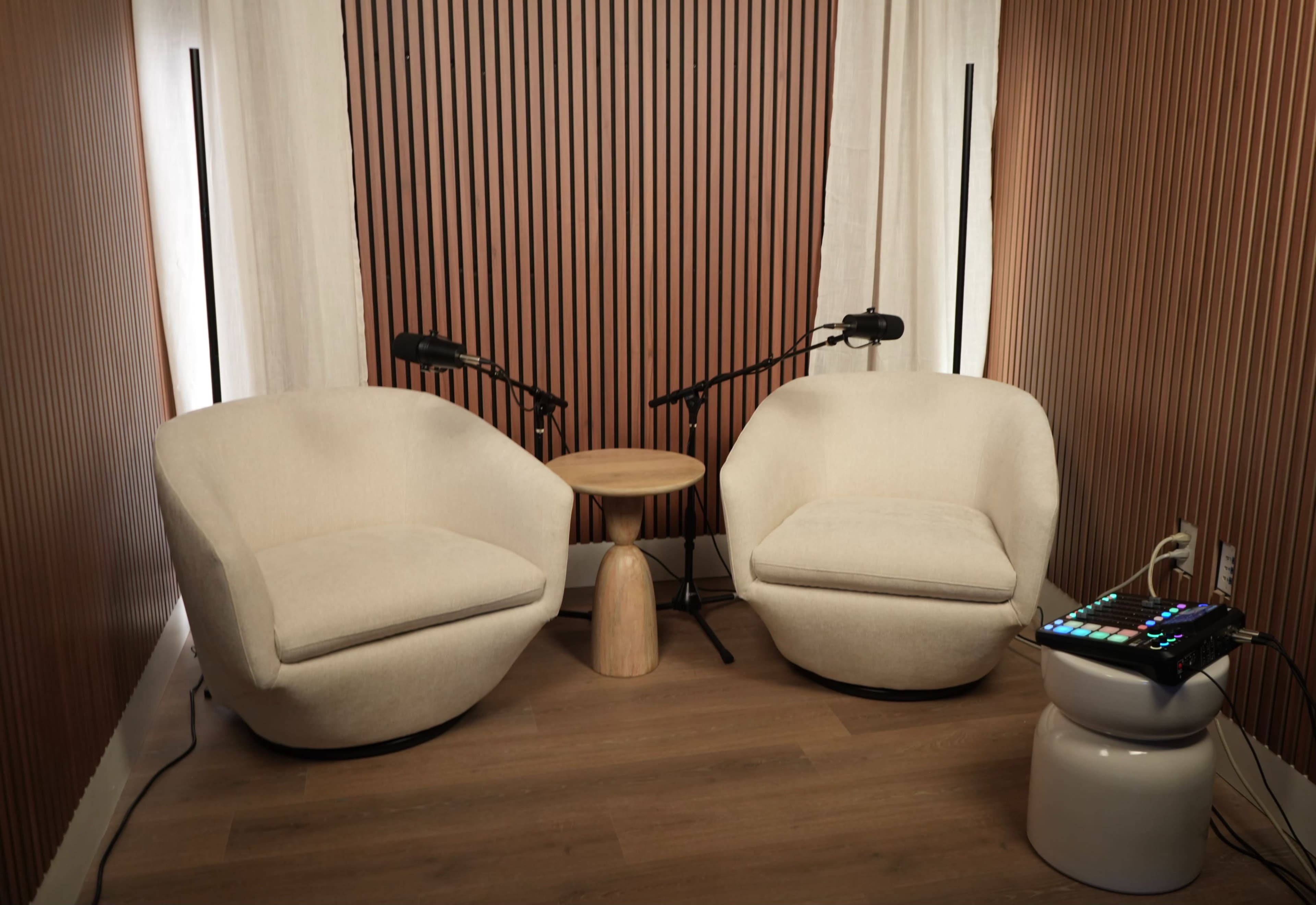 The image shows a cozy space with two round, cream-colored chairs, a wooden side table, and two microphones positioned on stands.