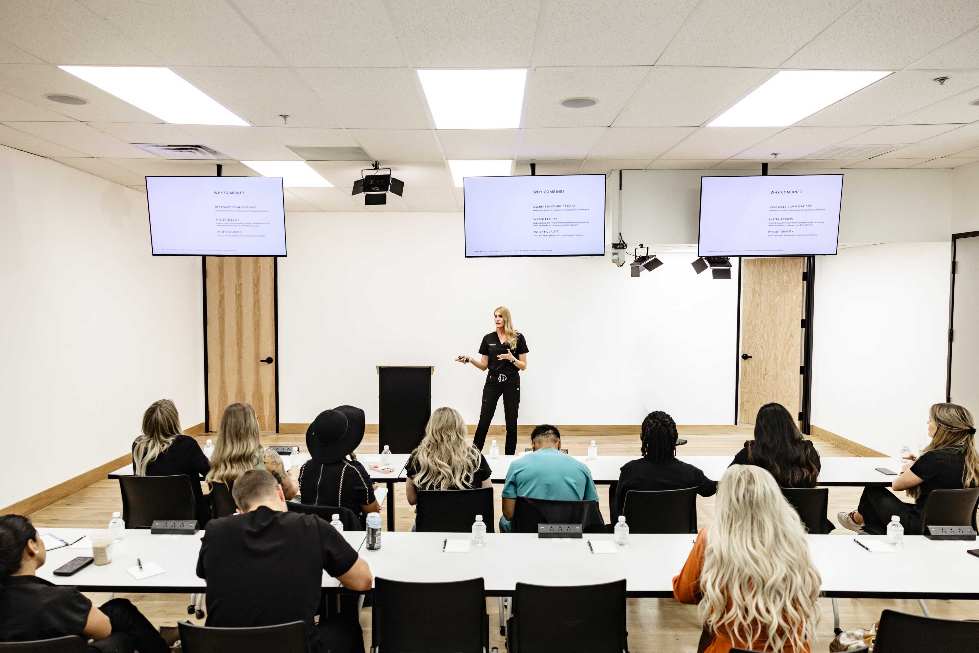 A woman stands in front of a classroom, presenting to a group of seated students while two screens display information above her.