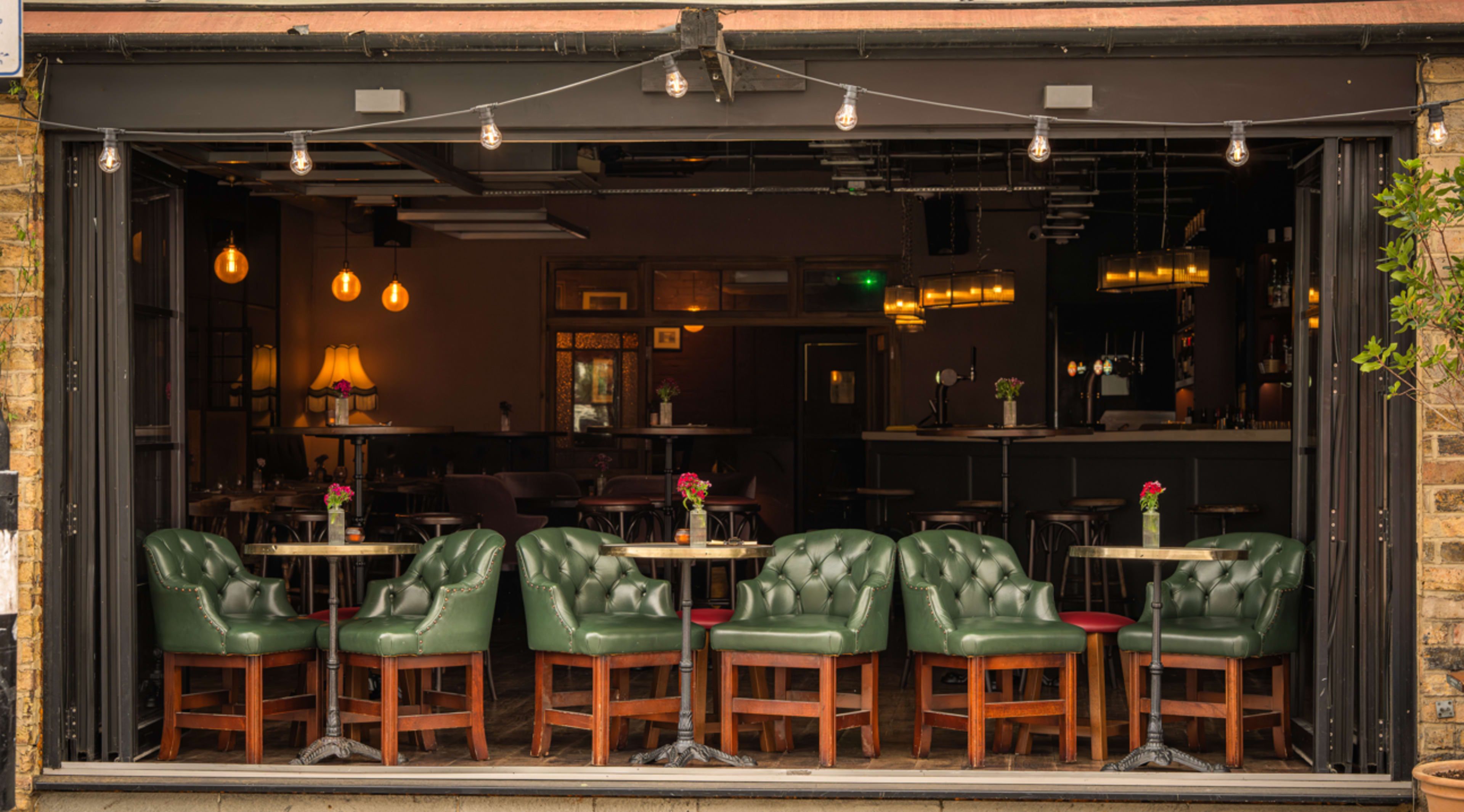 The image shows a restaurant interior with green upholstered chairs arranged around small tables, visible through an open facade featuring string lights above.