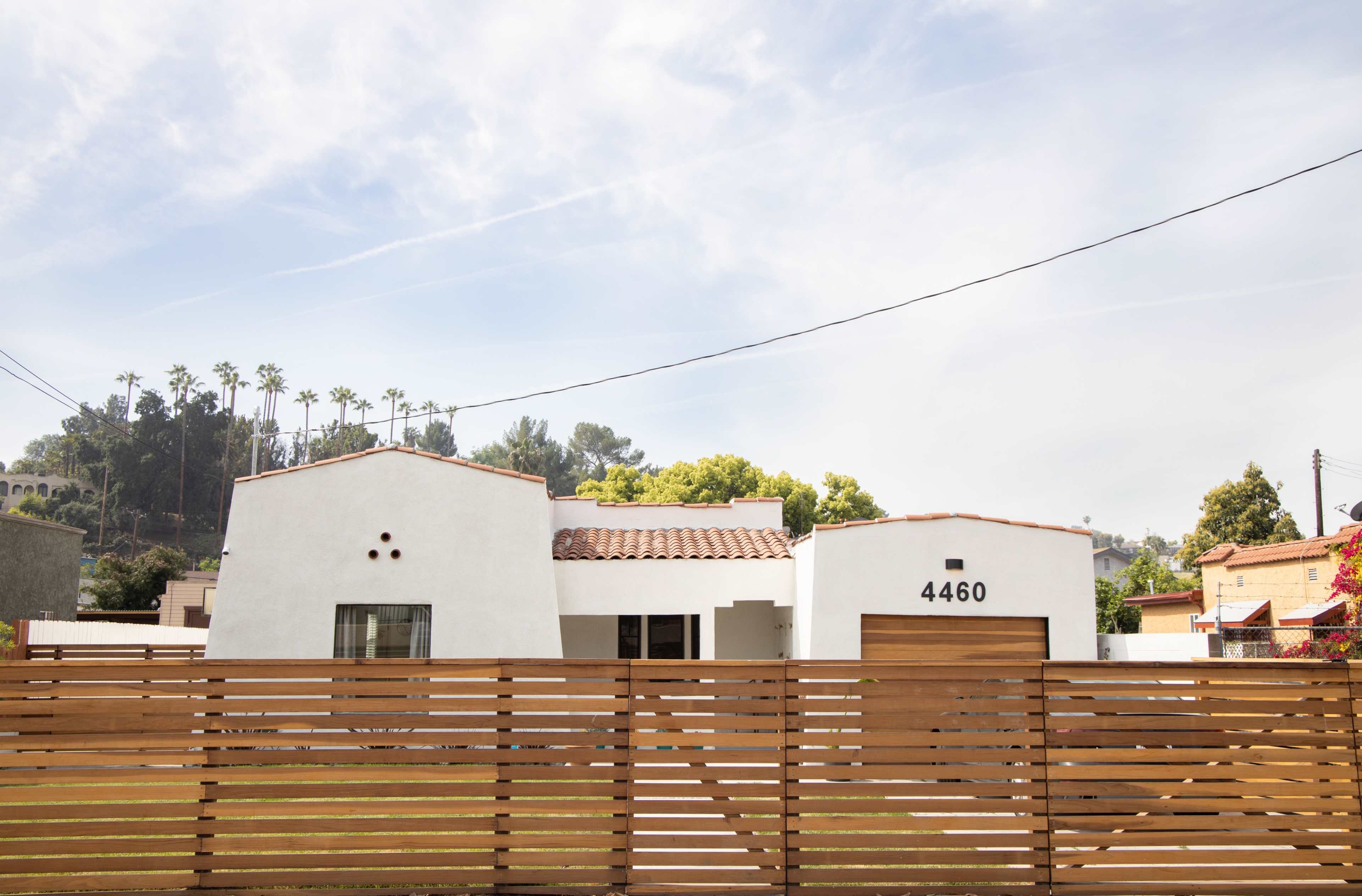 The image shows a modern, white house with a tile roof and a wooden fence, set against a backdrop of palm trees and hills.