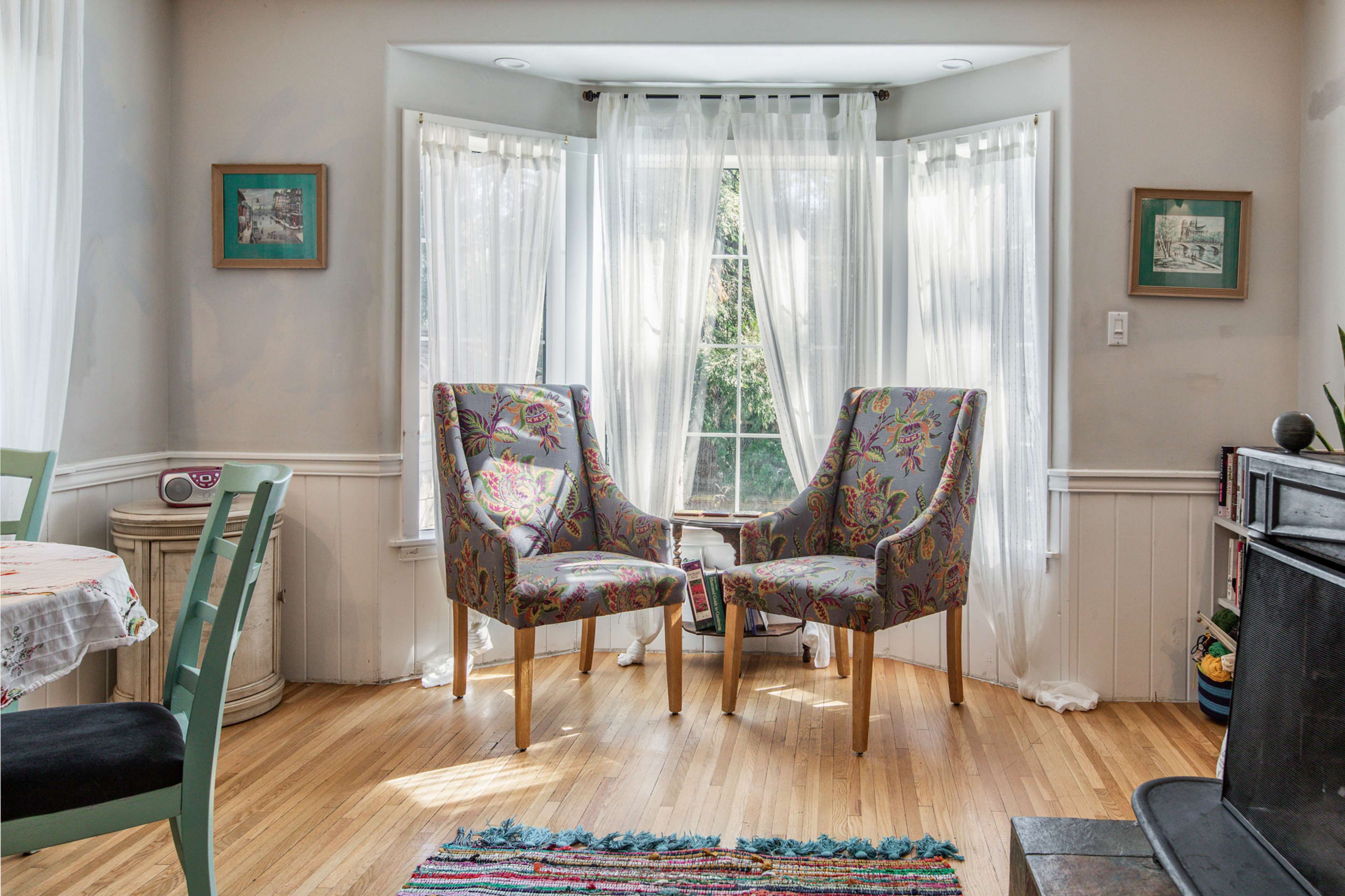A bright corner of a room features two floral-patterned chairs facing each other near a large window, with light curtains allowing natural light to filter in.