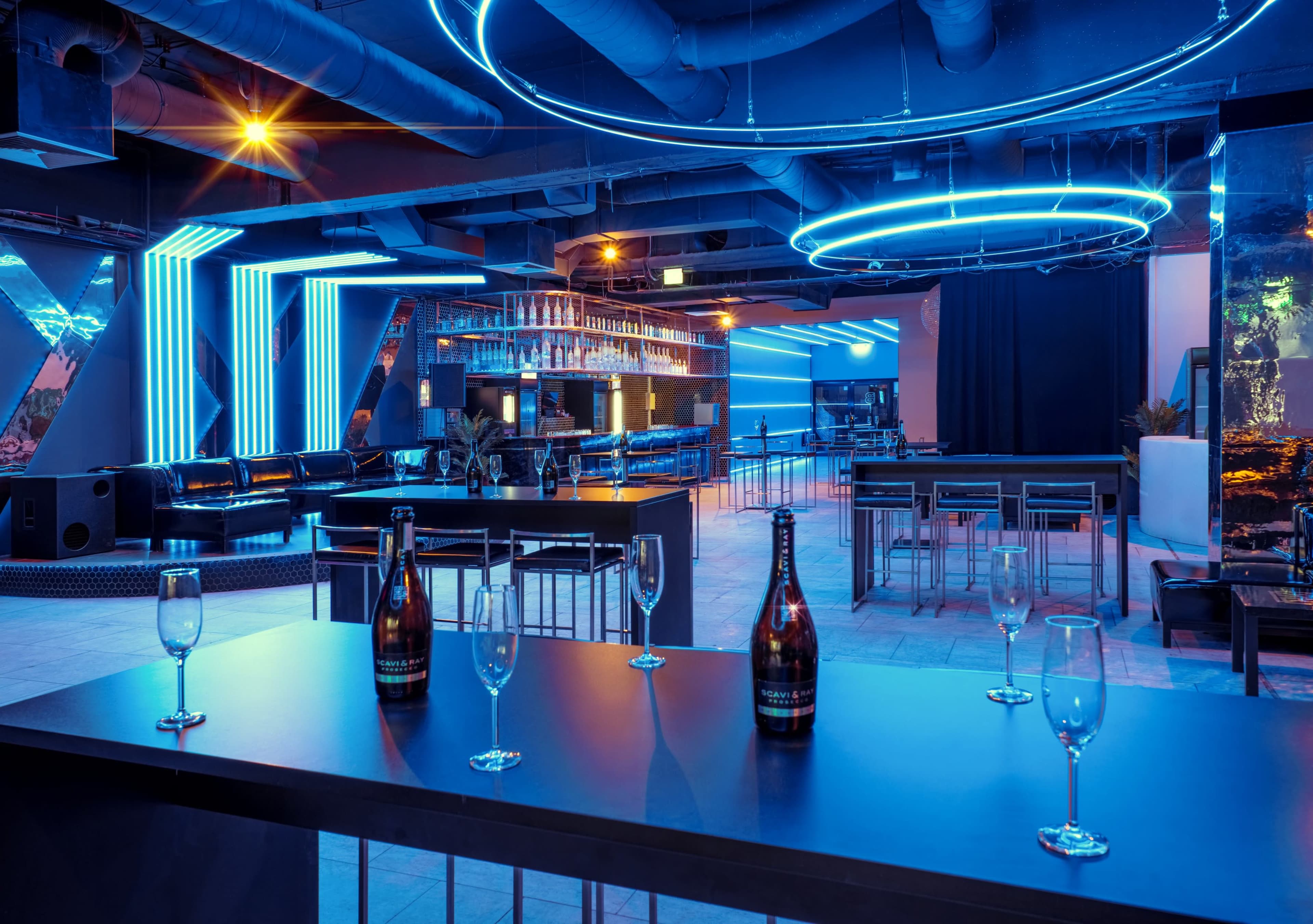 A modern bar interior featuring blue neon lighting, sleek furniture, and several empty champagne flutes on a black countertop.