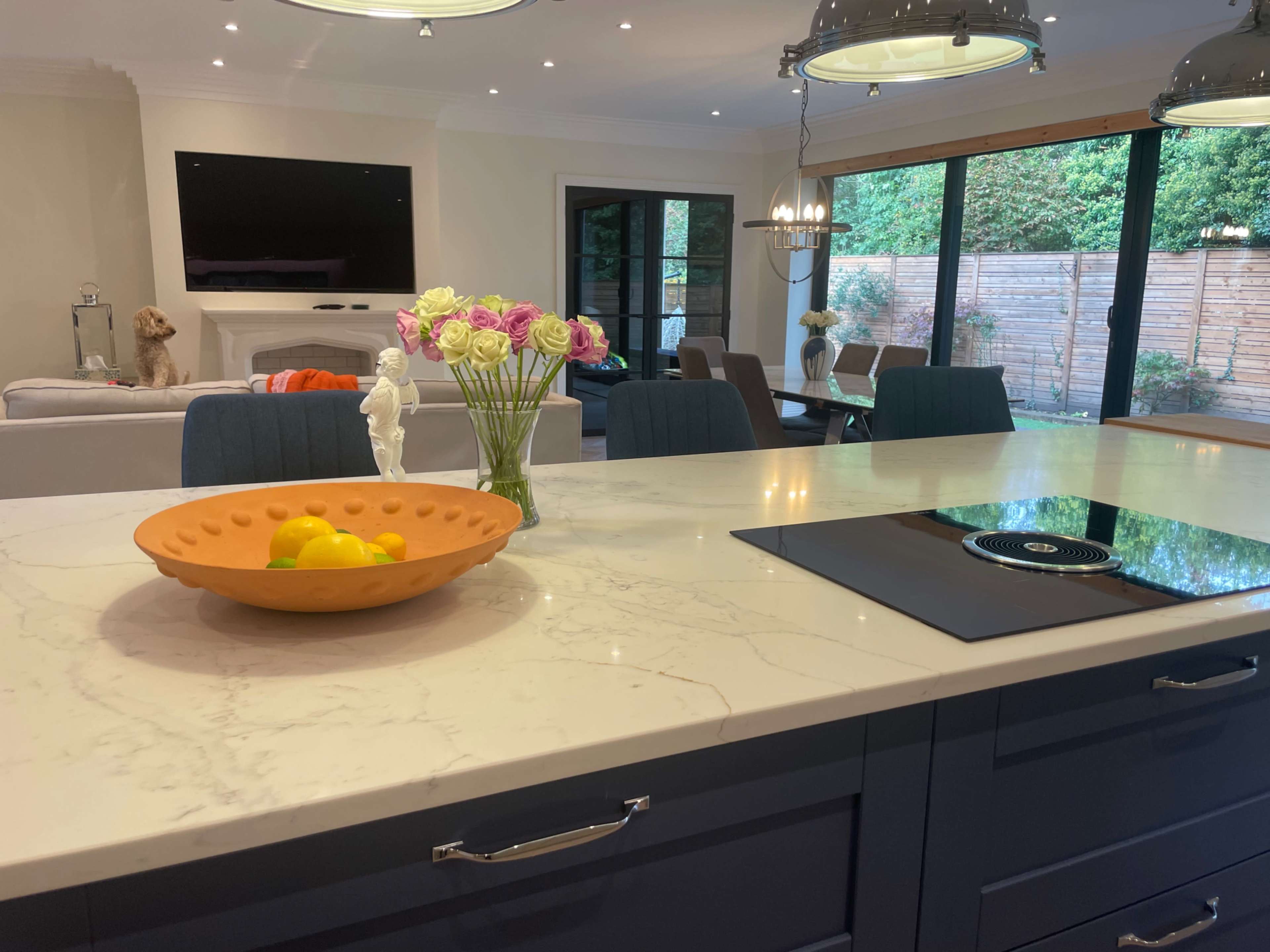 The image shows a modern kitchen with a marble countertop featuring a bowl of lemons, along with a view of a living area and outdoor garden through large glass doors.