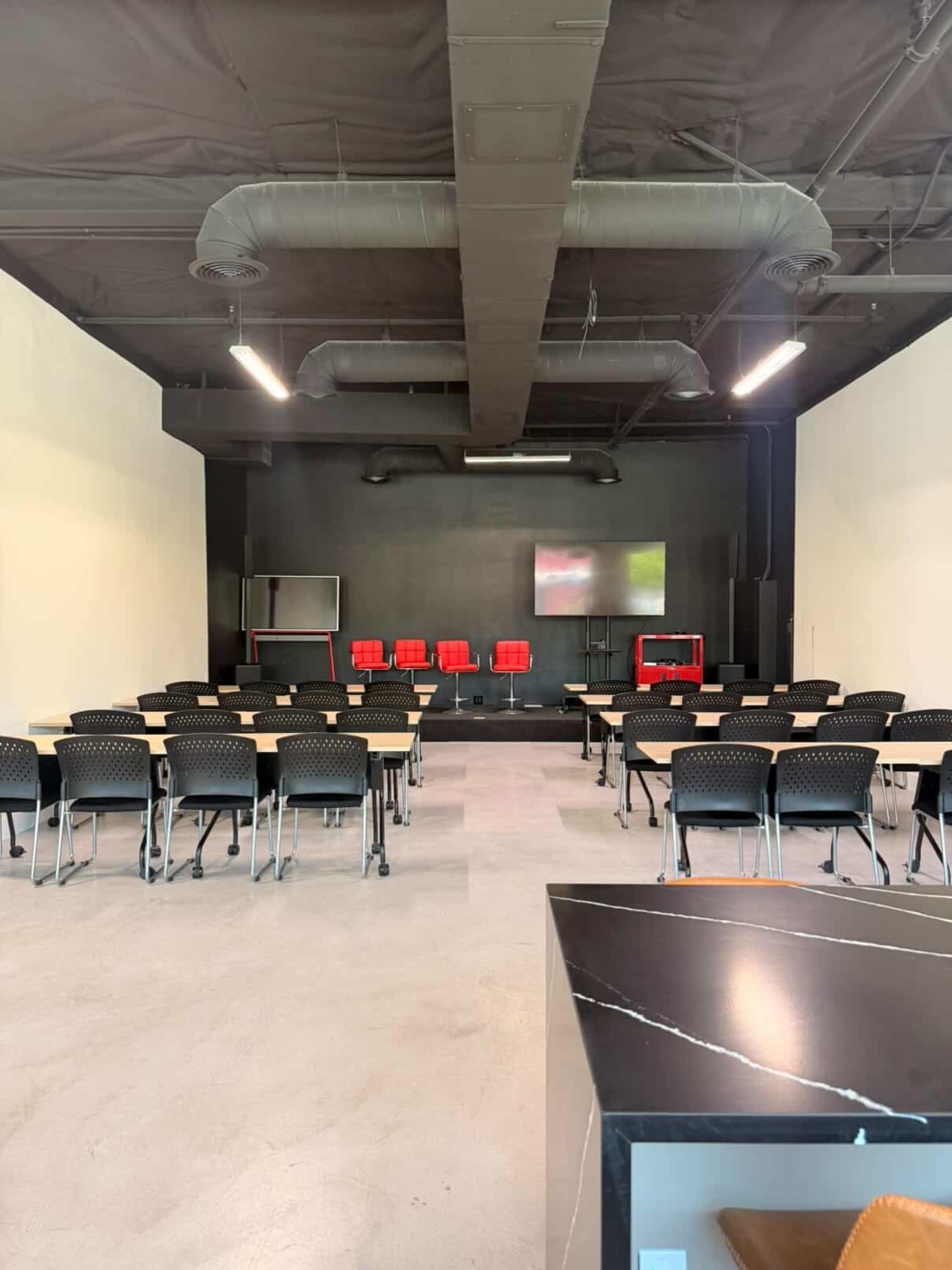 The image shows a conference room with rows of black chairs and tables facing a screen, along with a few red chairs positioned at the front.
