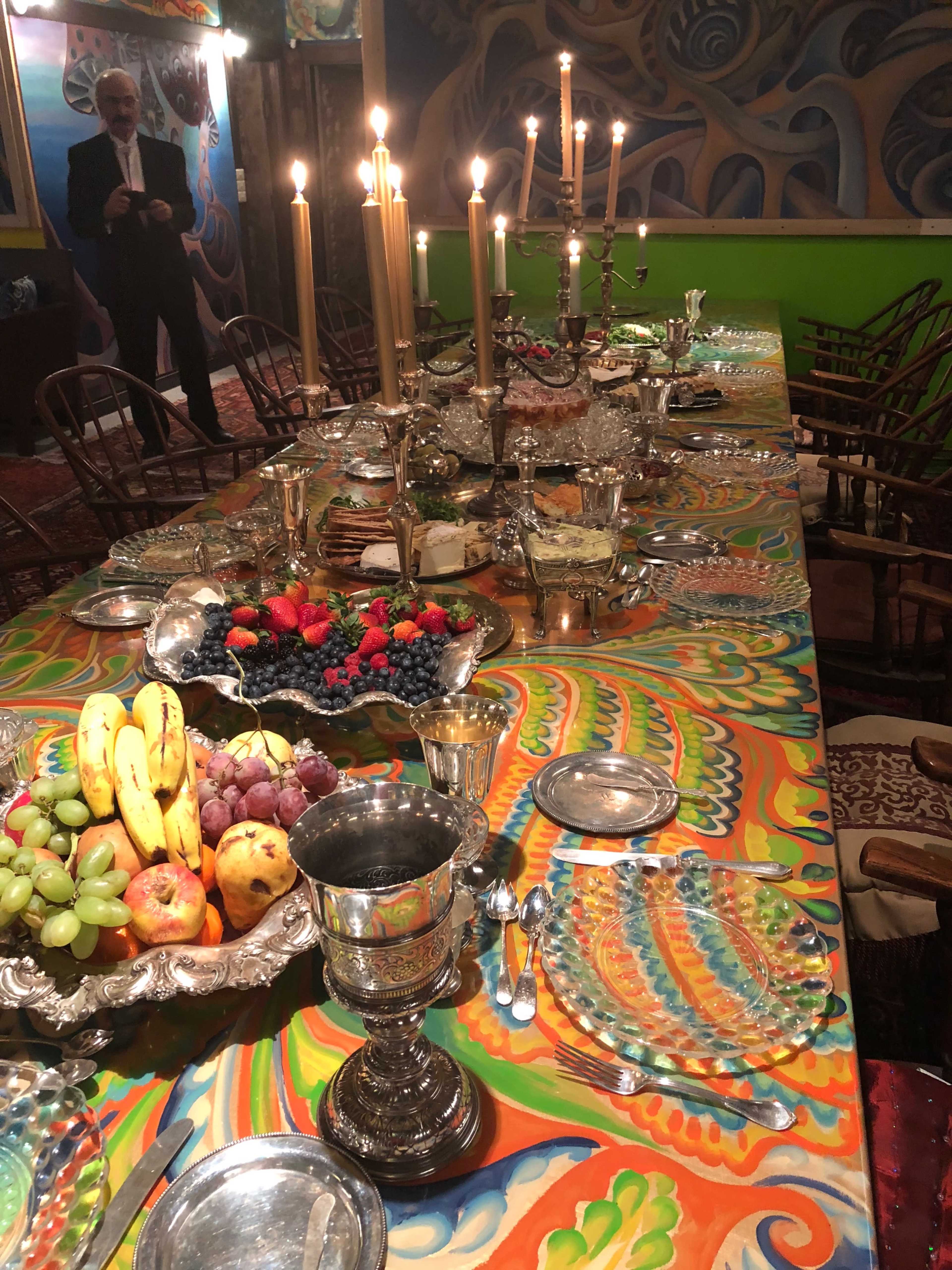 A long table set with an array of food, silverware, and decorative candles, surrounded by vibrant artwork and a figure standing in the background.