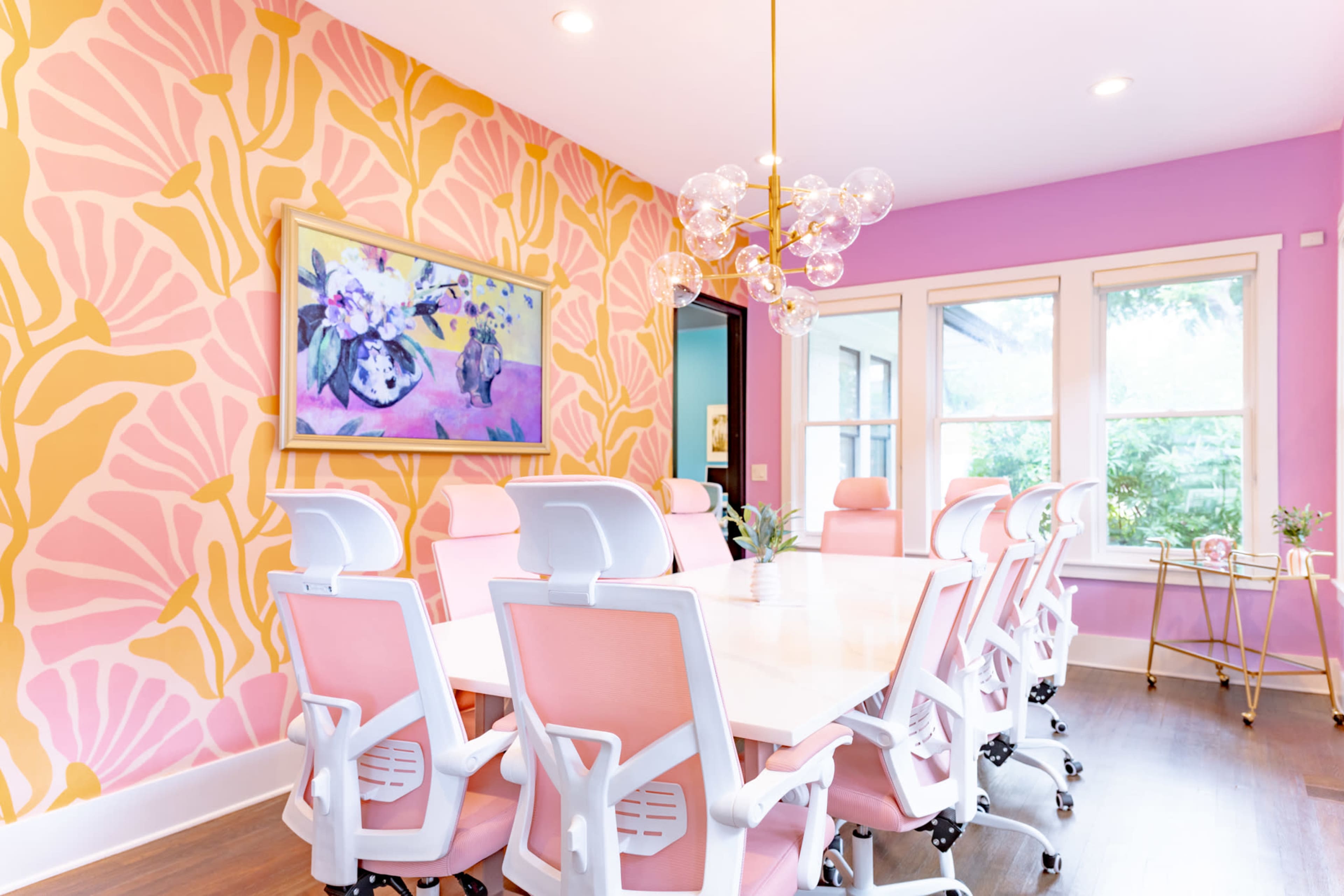 A brightly colored conference room features a large table surrounded by pink ergonomic chairs against a decorative floral wallpaper.