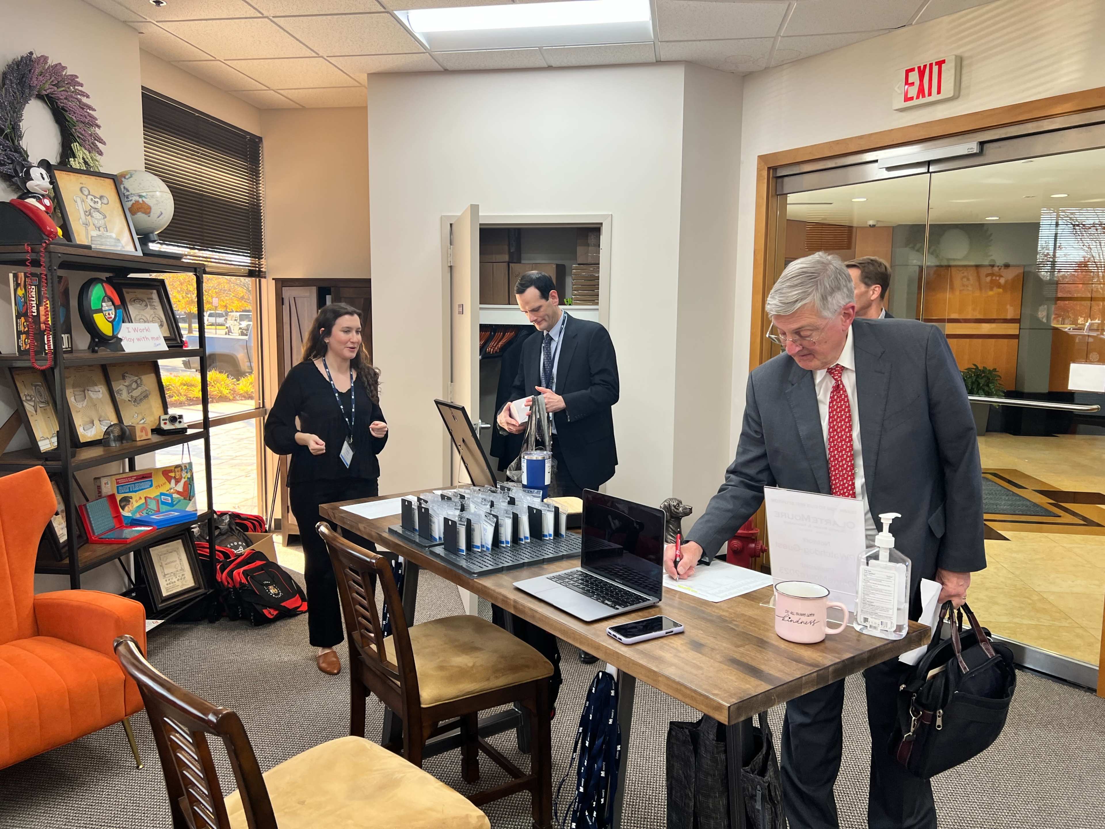 A group of professionals gathers at a table in a reception area, with documents and a laptop visible on the surface.