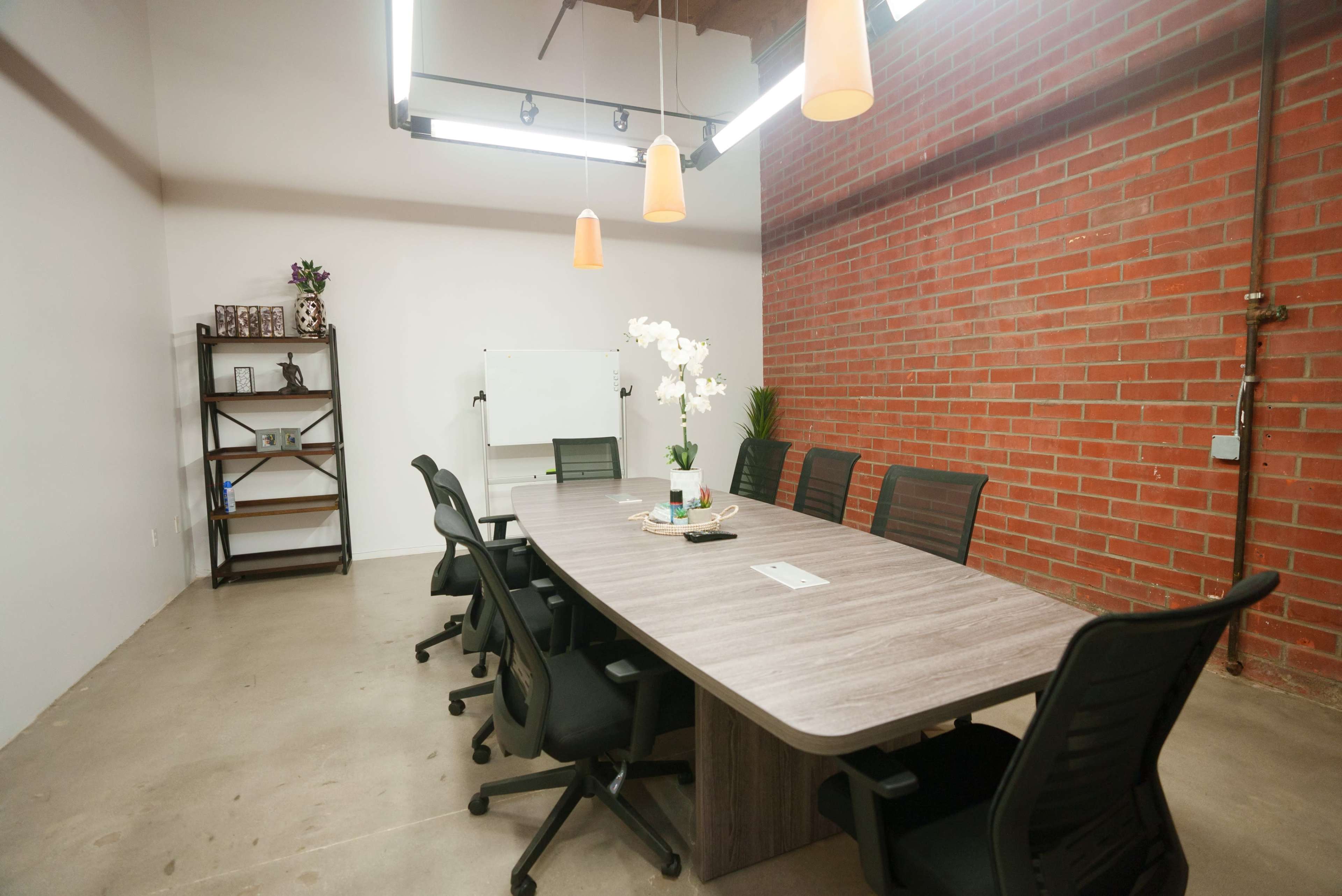 A modern conference room features a long wooden table surrounded by black chairs, with a brick wall and overhead lighting.