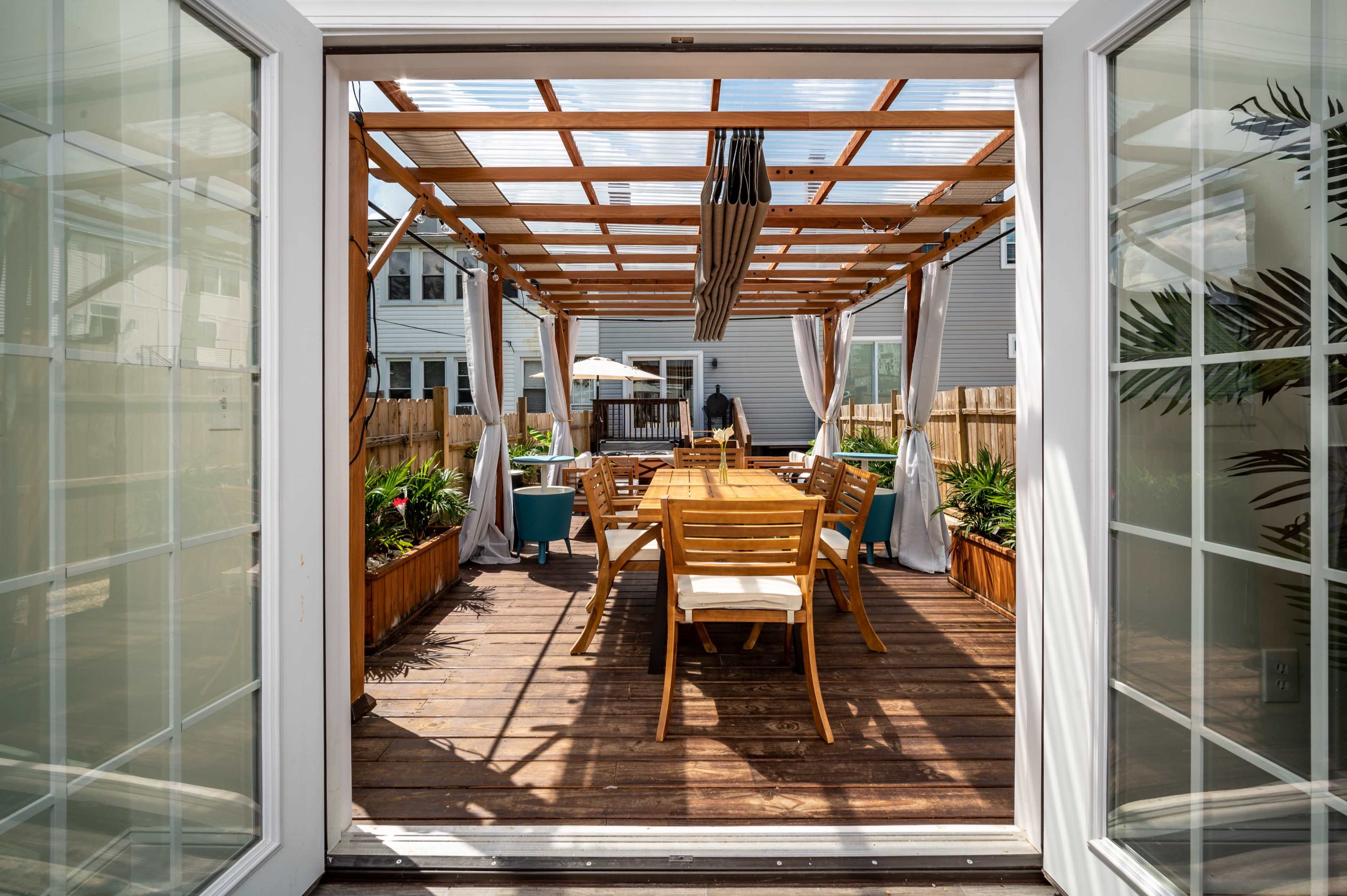 A wooden deck with a table and chairs is partially enclosed by a pergola, visible through open double doors.