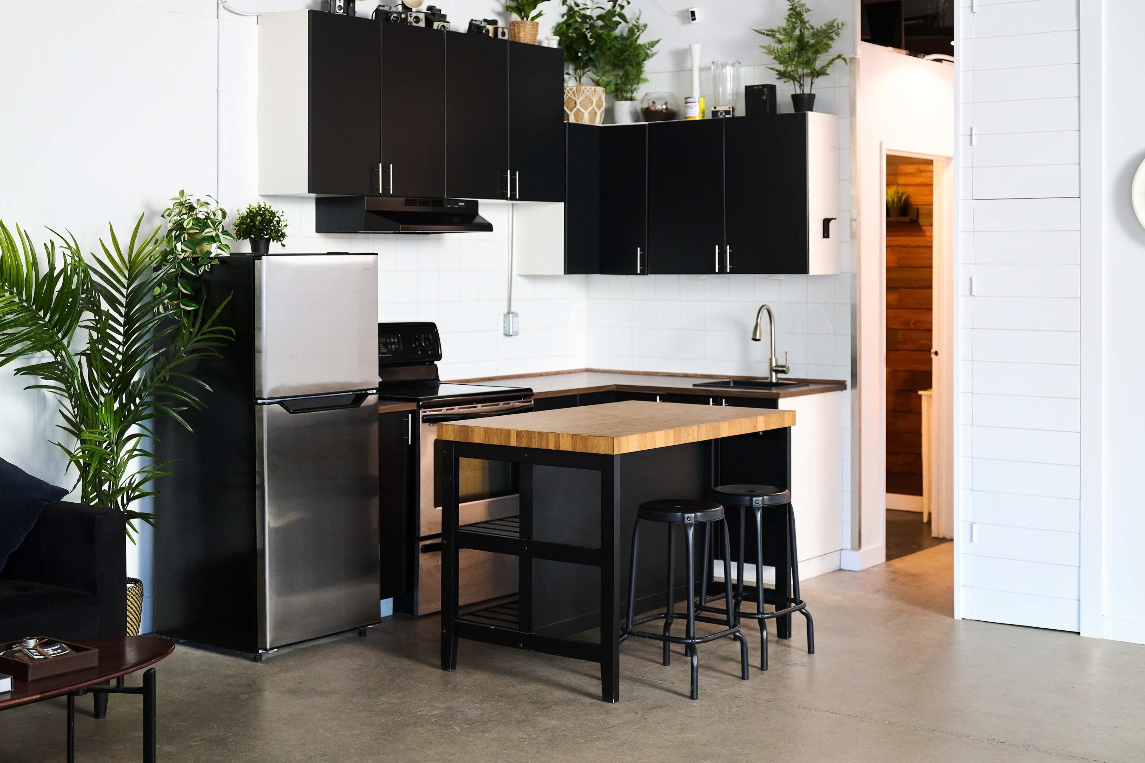 The image shows a modern kitchen with black cabinets, stainless steel appliances, a wooden countertop island, and two black stools.