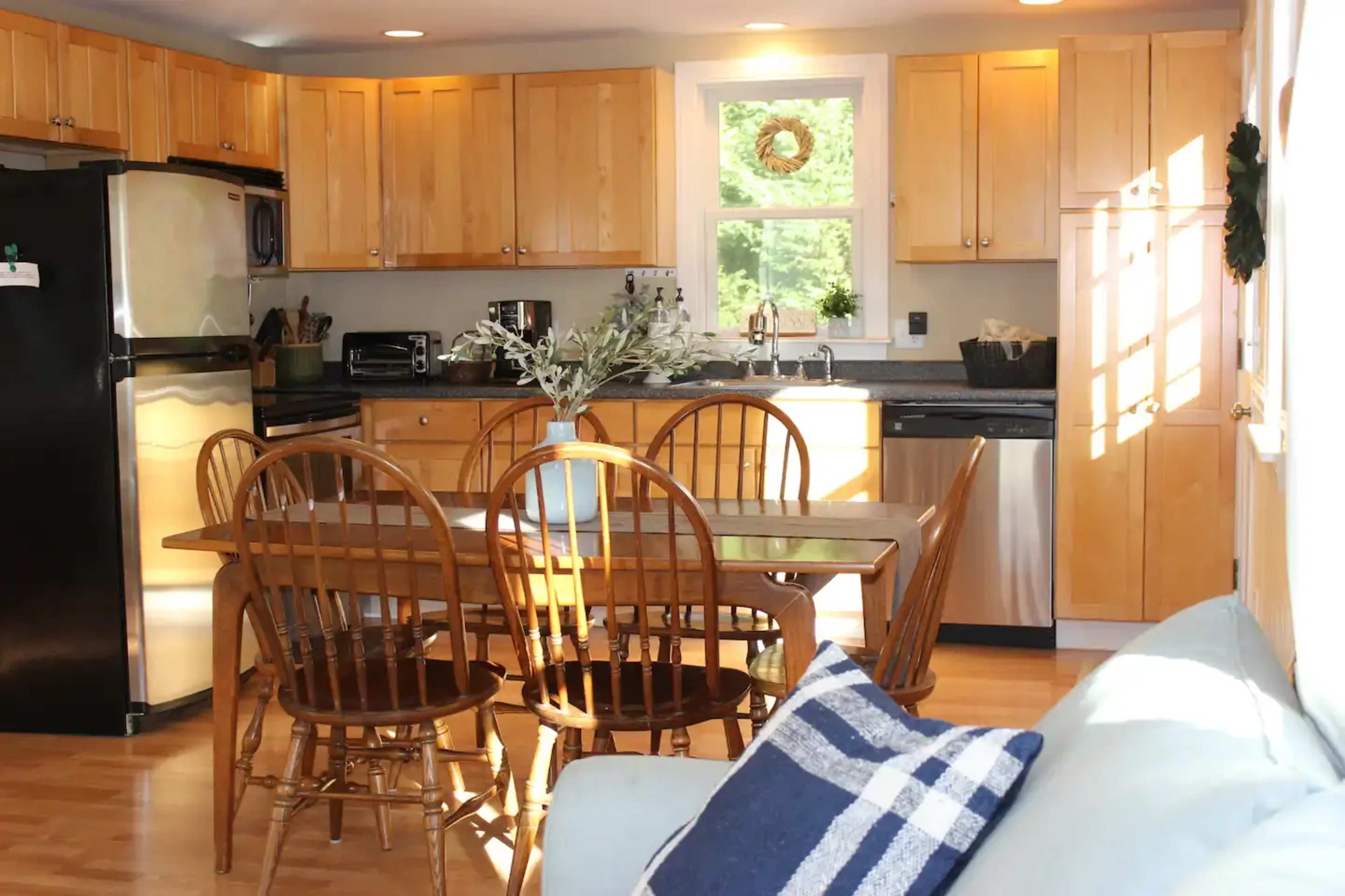 A kitchen with wooden cabinets, a black refrigerator, and a dining table surrounded by four wooden chairs, with a couch visible in the foreground.