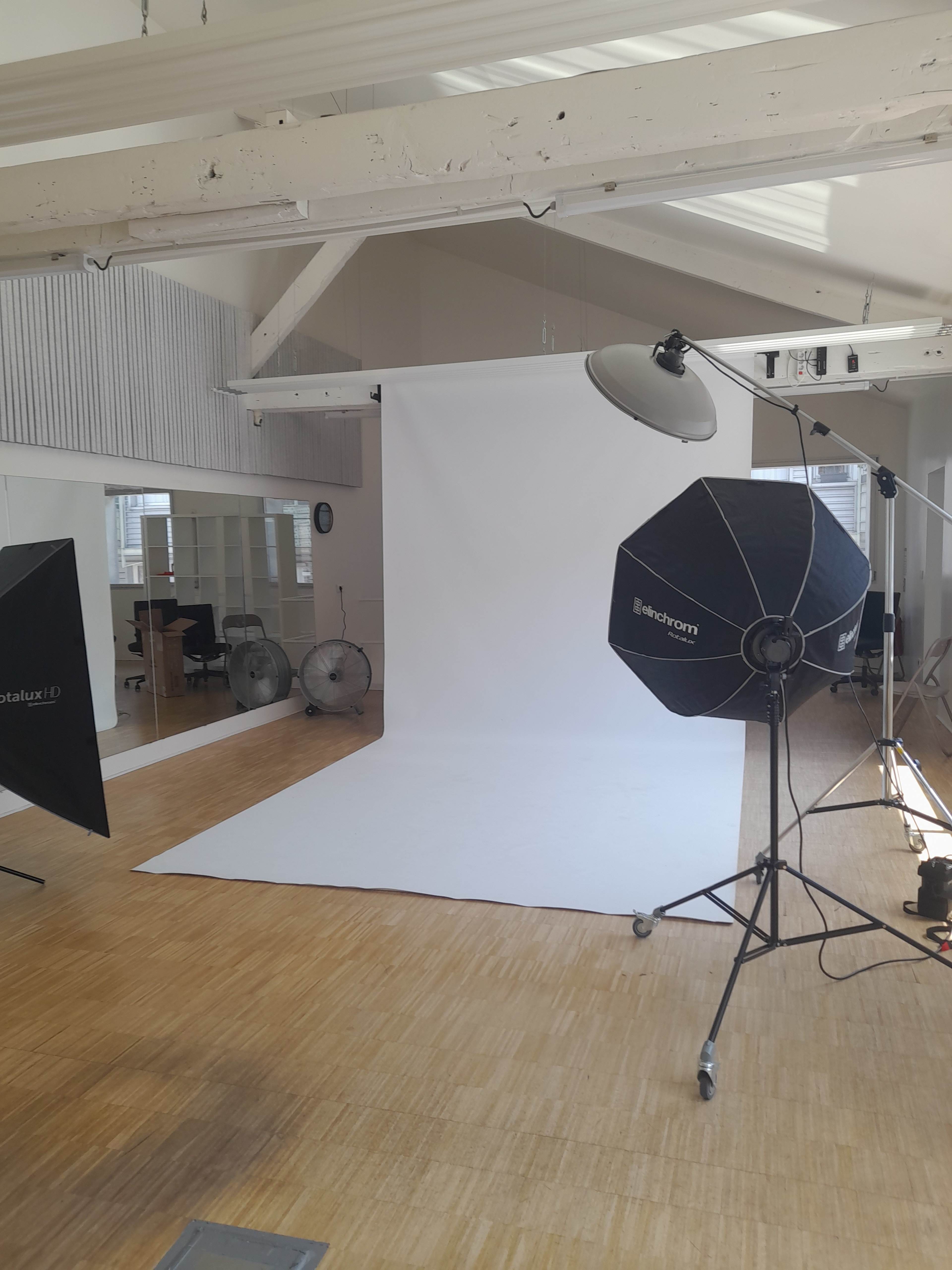 The image shows a spacious photography studio with a white backdrop, lighting equipment, and fans arranged on wooden flooring.