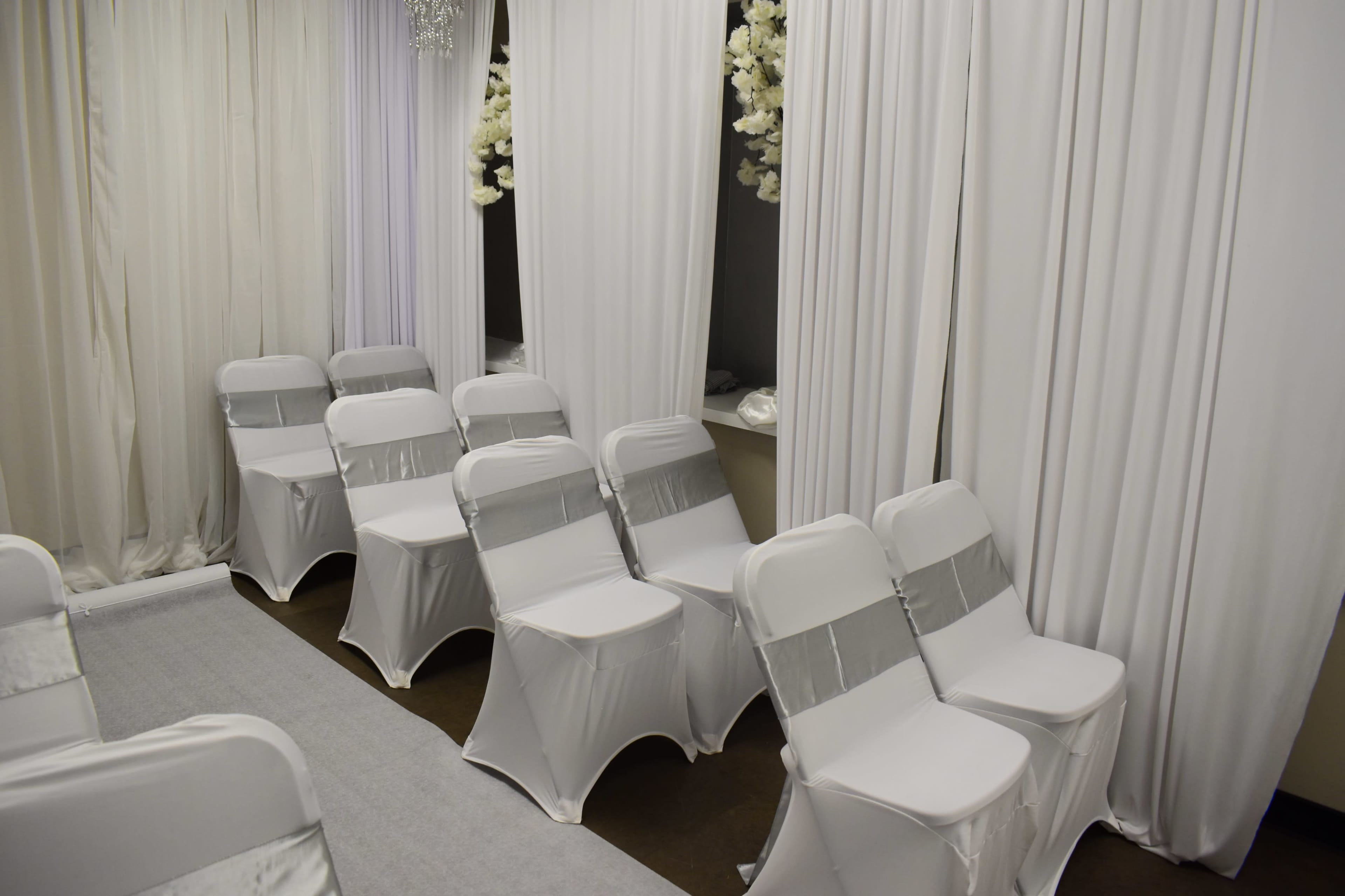 The image shows a row of white chairs with silver sashes arranged neatly in a softly lit space decorated with white drapes and floral accents.