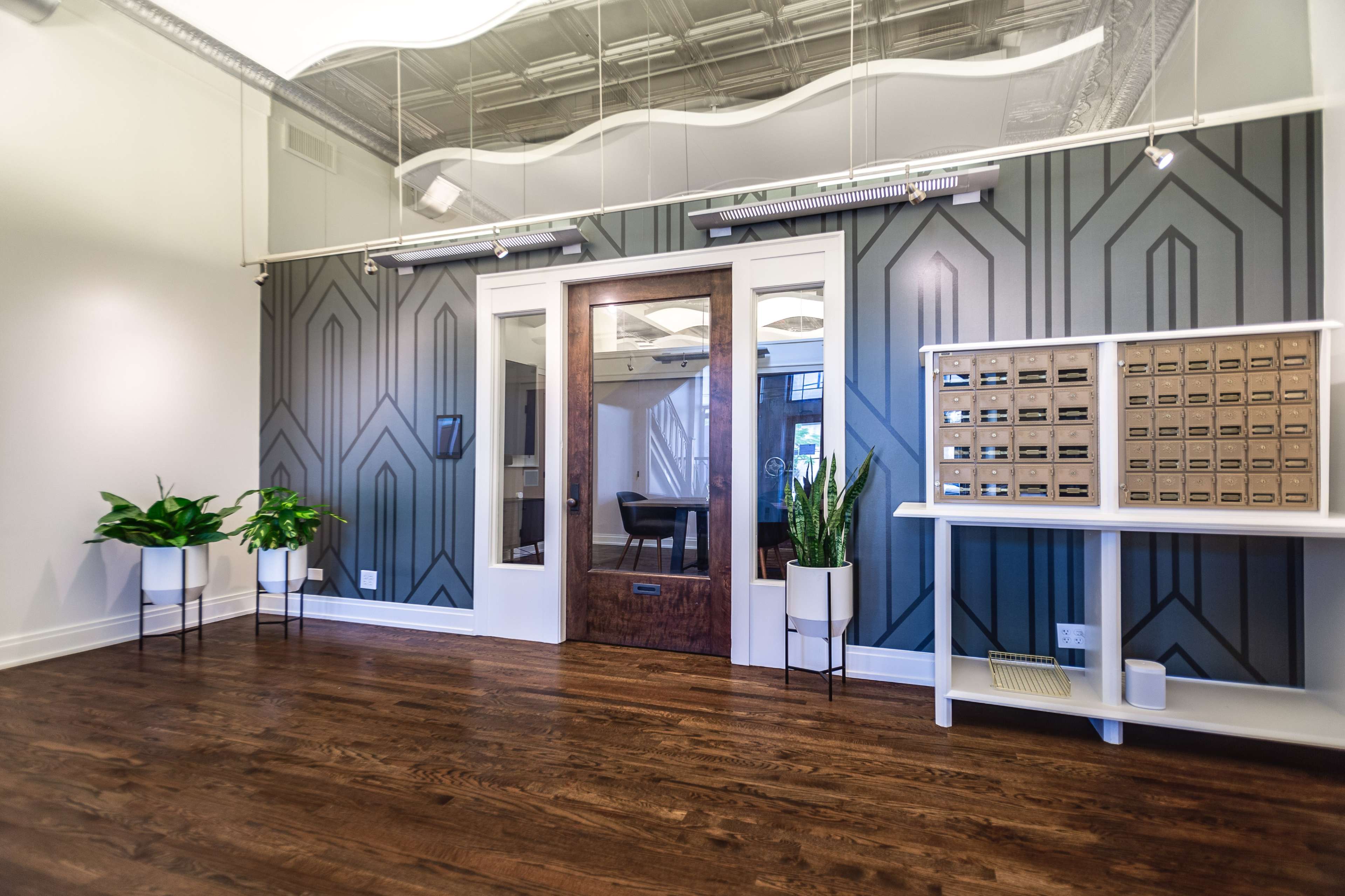 A modern interior office space featuring a wooden door, decorative wall with geometric patterns, a row of plants, and a mailbox system on a white shelf.