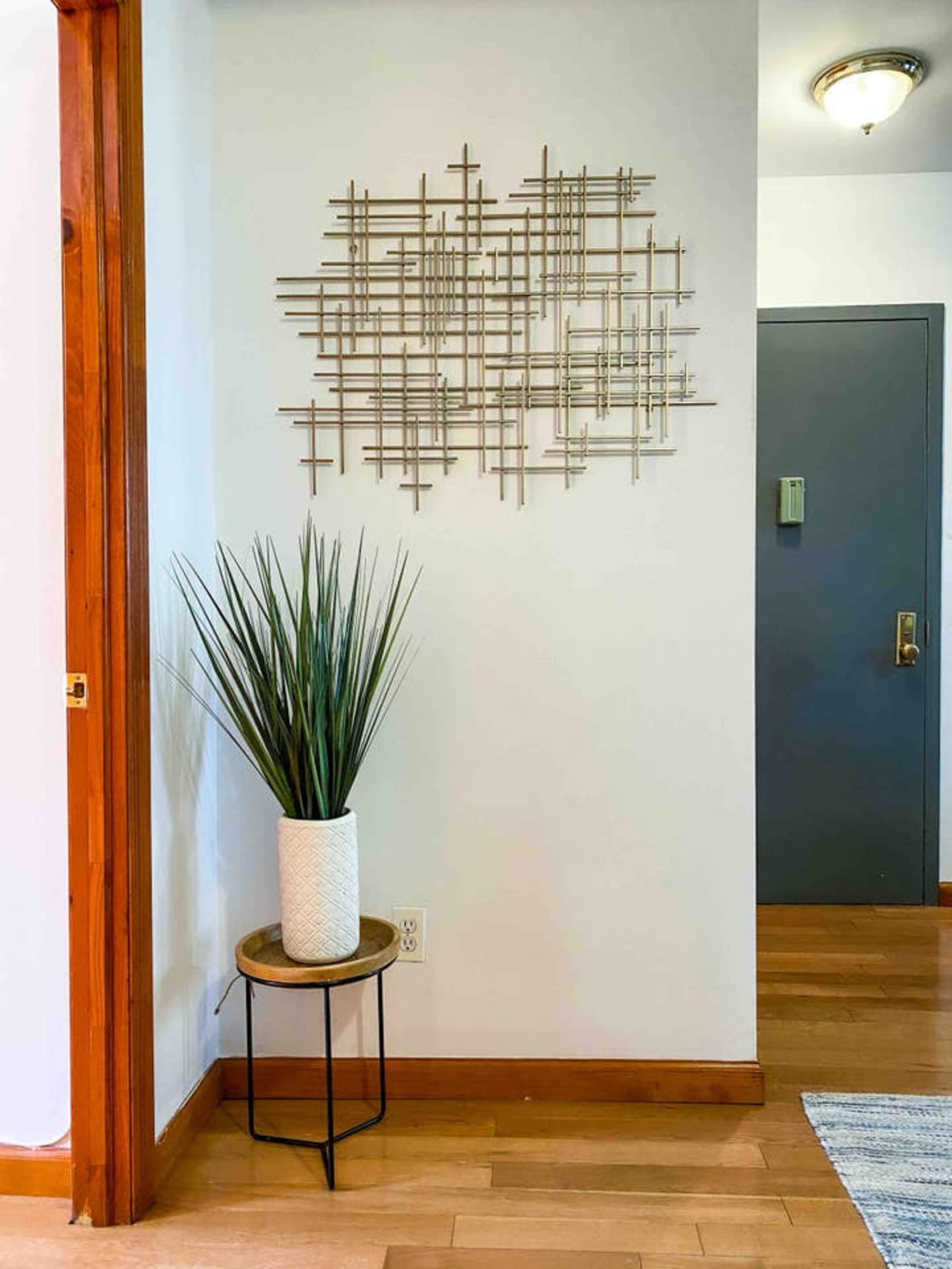 A decorative metal wall art piece is displayed on a white wall next to a potted plant on a small round table, with a closed door in the background.