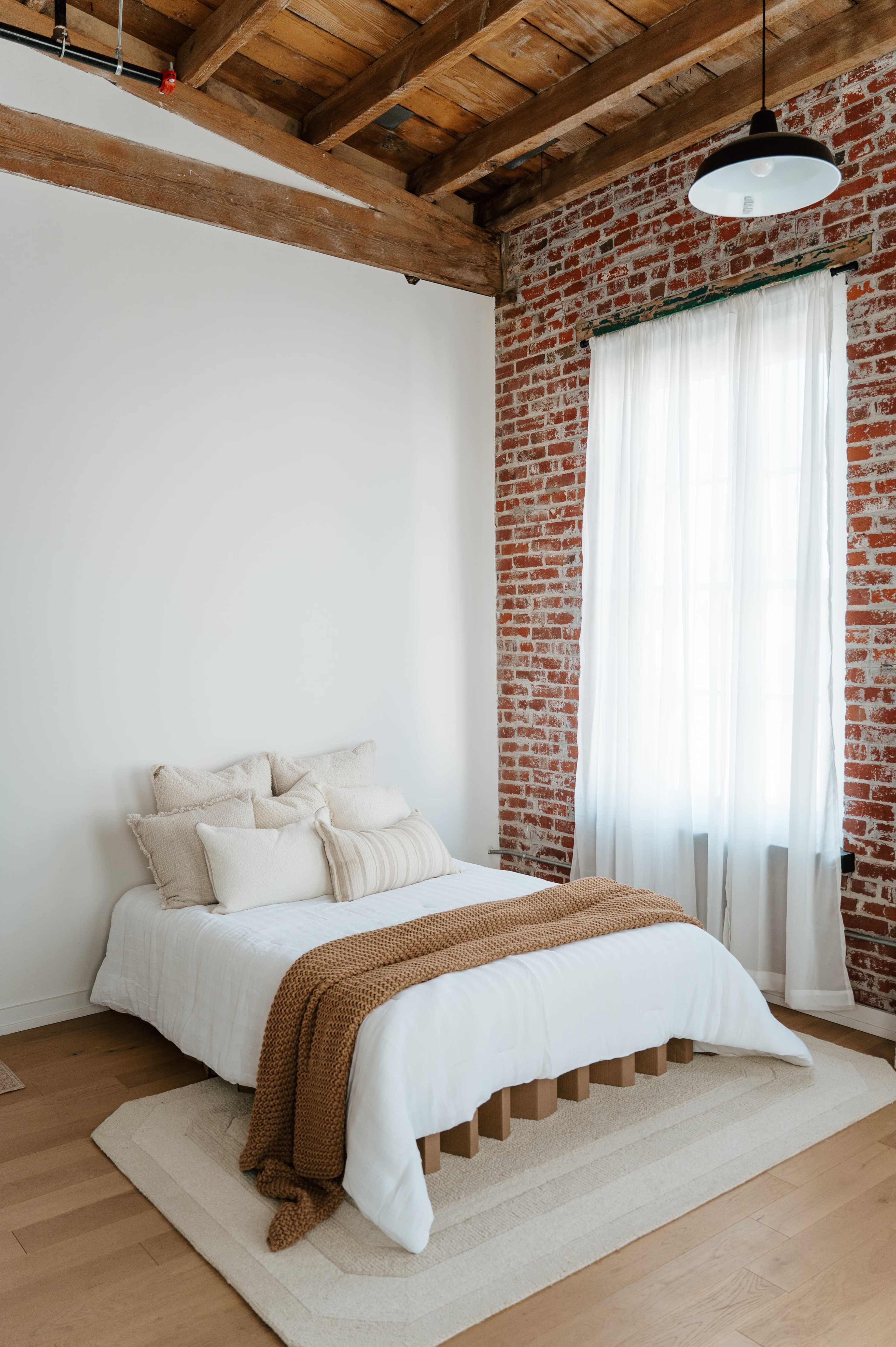 A neatly made bed with a light-colored blanket and decorative pillows is positioned in a room featuring exposed brick walls and a large window with sheer curtains.