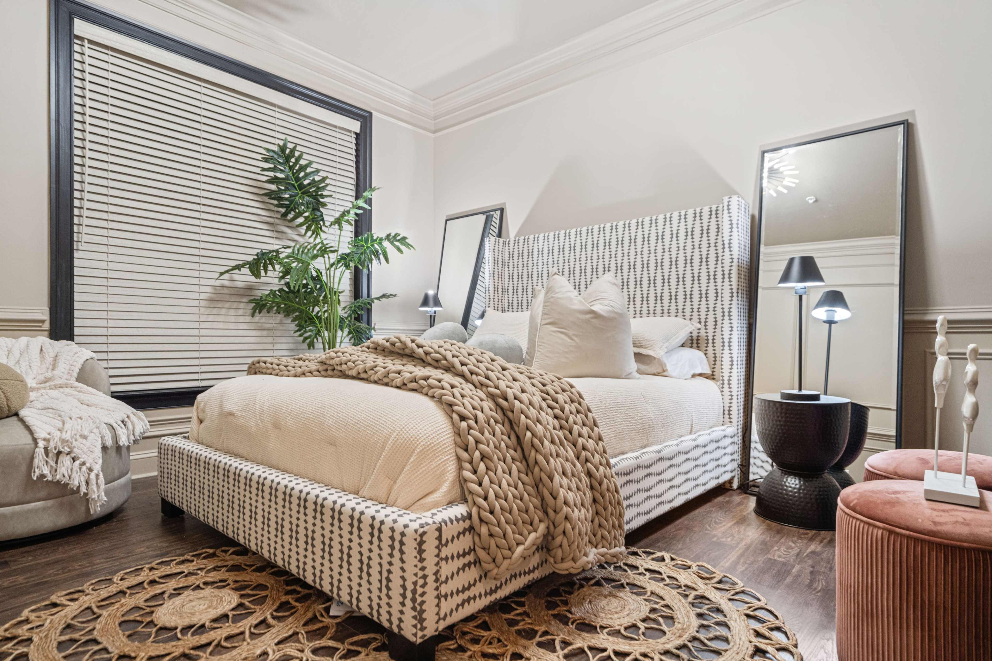 A modern bedroom featuring a patterned bed frame, a large woven throw blanket, two mirrors, a plant, and accent furniture.