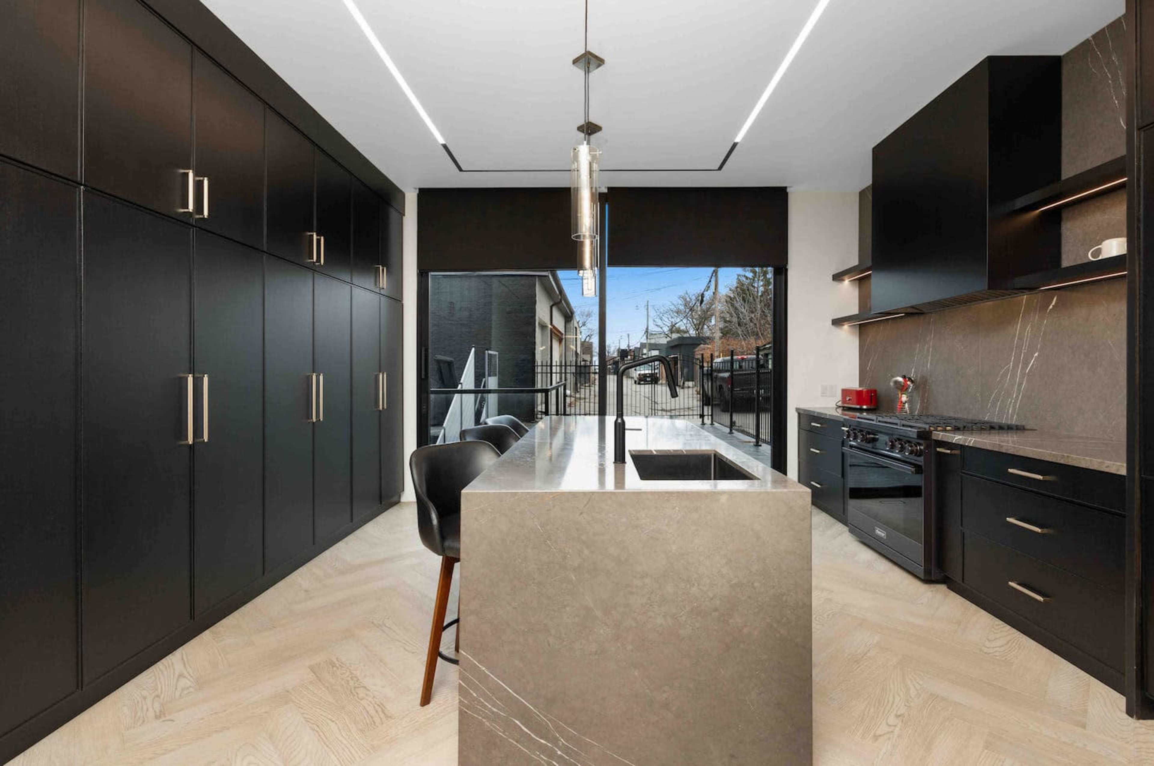 The image shows a modern kitchen featuring black cabinetry, a central island with a sink, and large windows that provide outdoor views.
