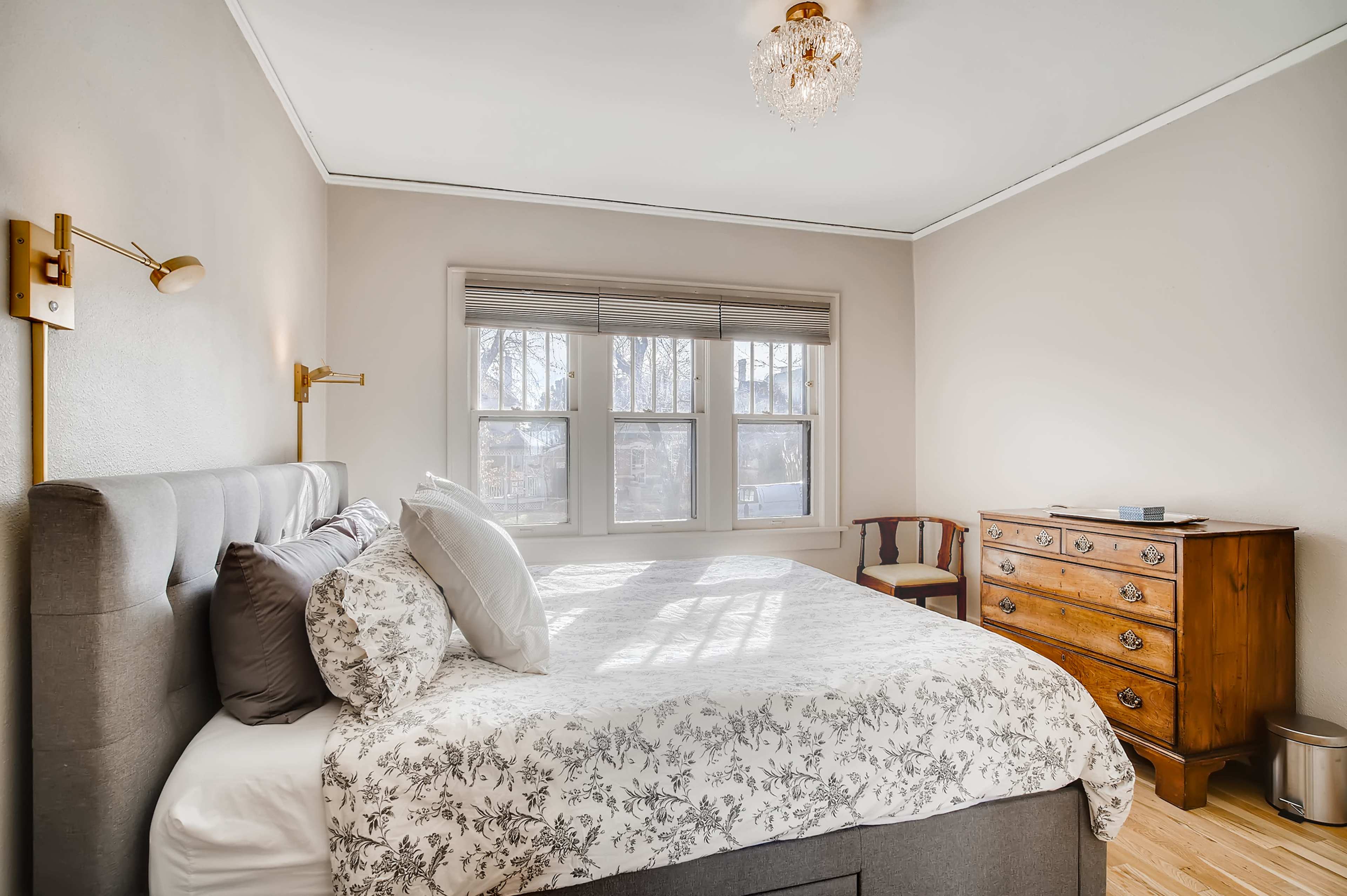 The image shows a cozy bedroom with a gray upholstered bed, a wooden dresser, and a chair positioned near a window.