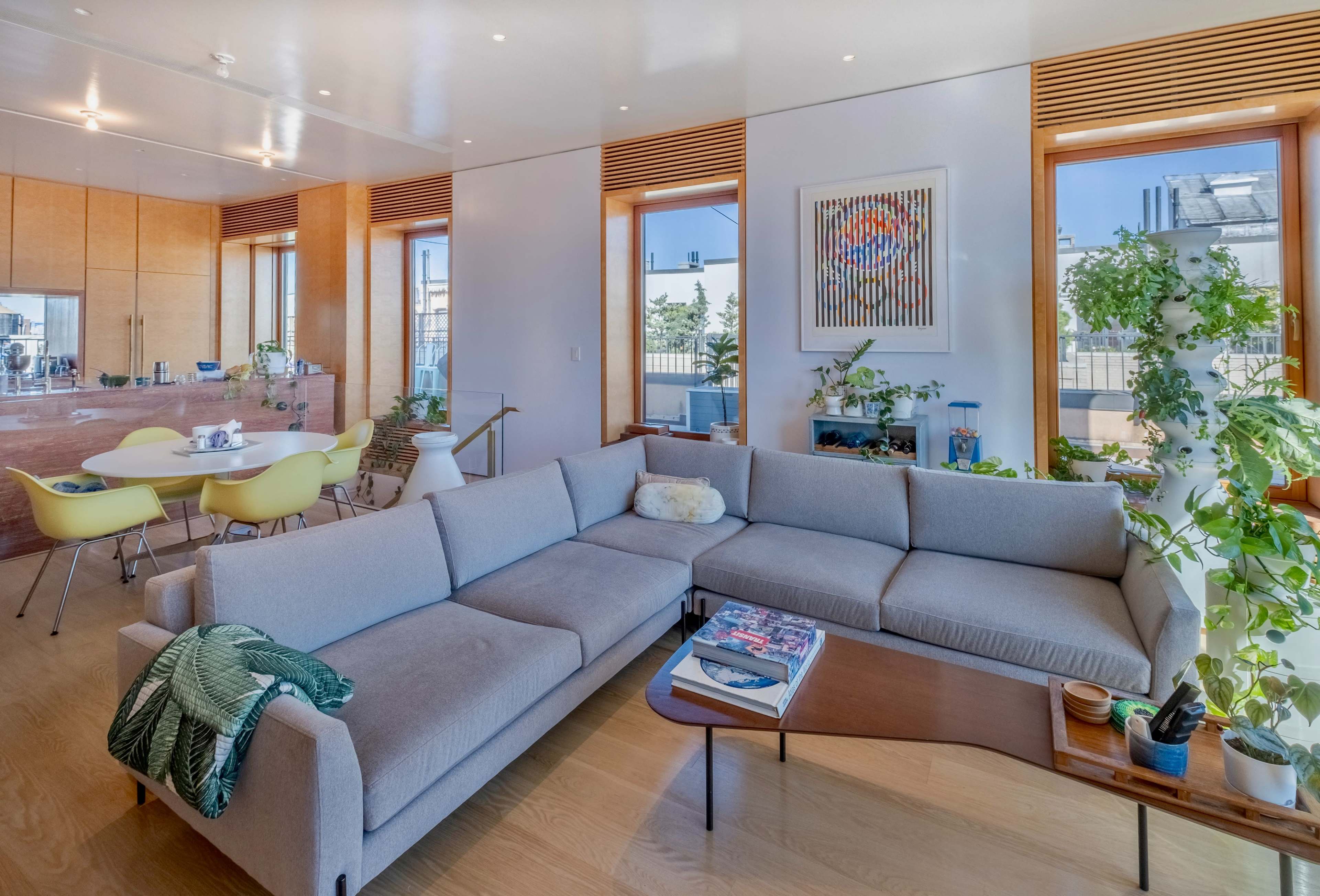 A modern living room featuring a gray sectional sofa, a wooden coffee table, large windows with plants, and a dining area in the background.