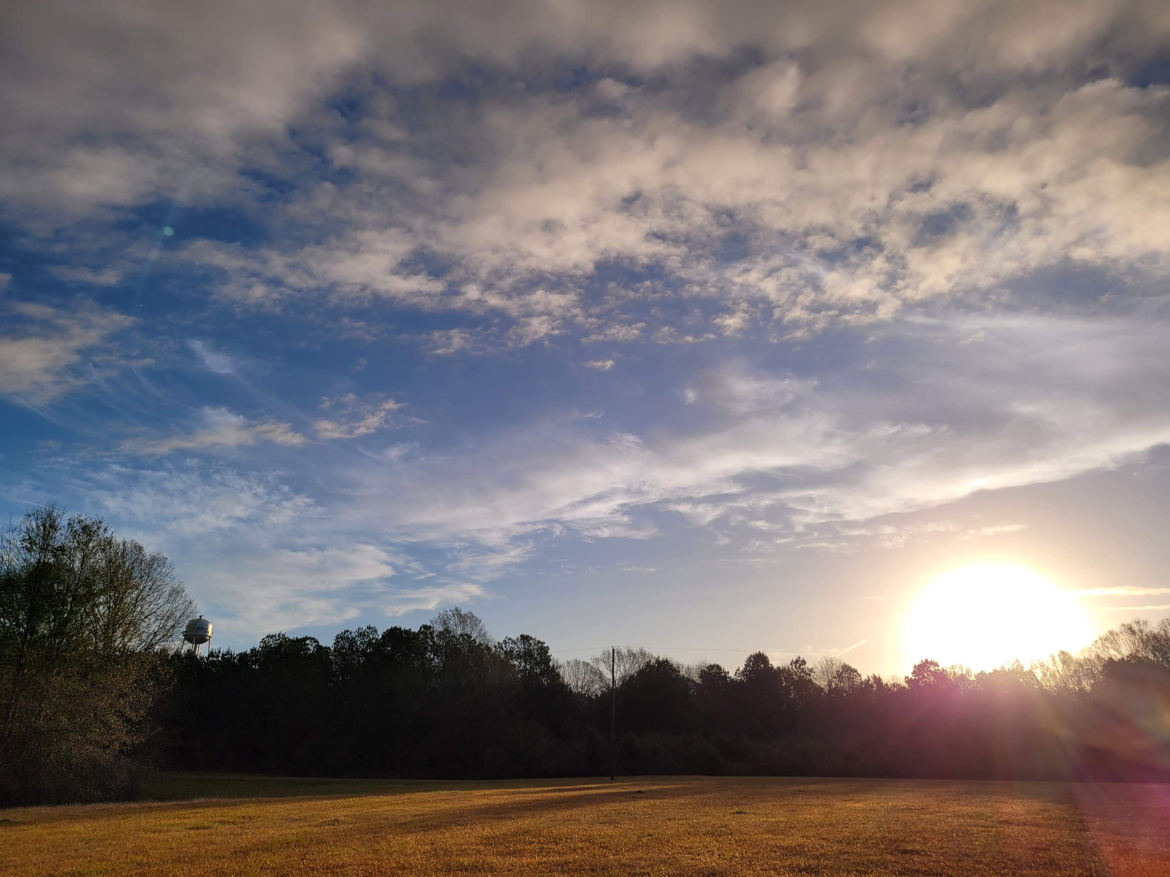 The image shows a sun setting behind a grassy field with trees and a water tower in the background under a partly cloudy sky.