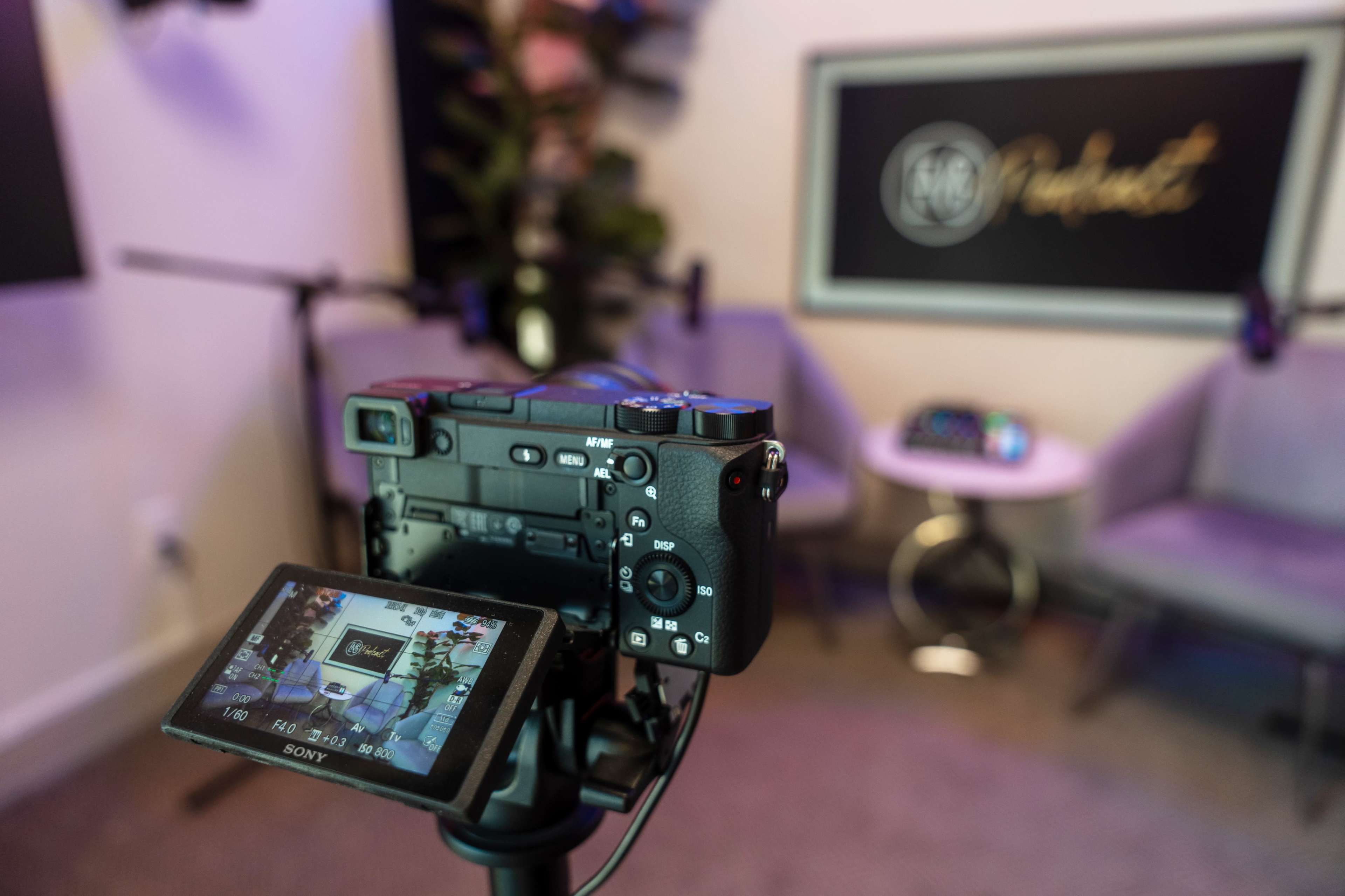 A Sony camera is positioned on a tripod, focused on a setup for a podcast recording, featuring a microphone and a small table with chairs.