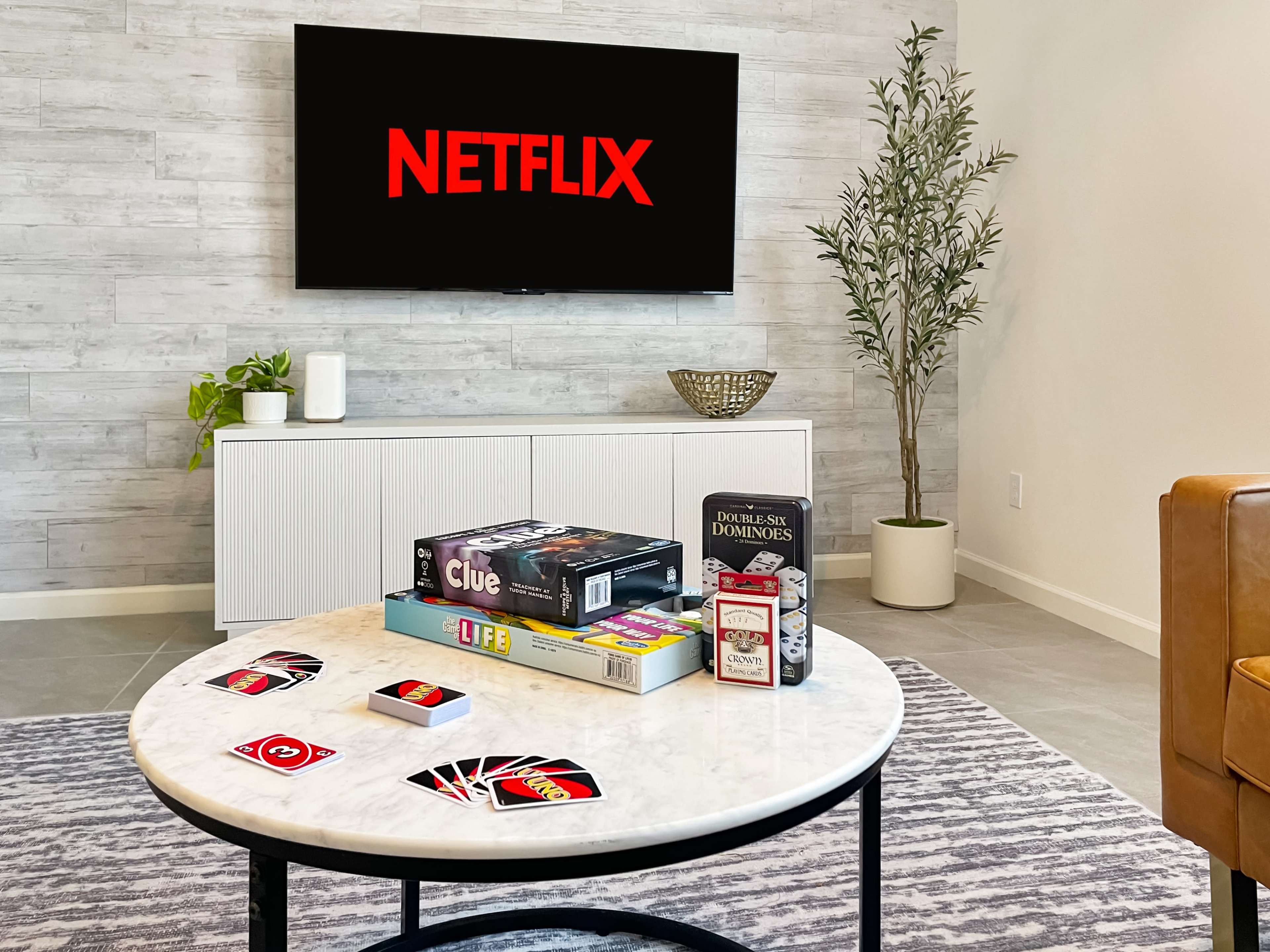 A modern living room features a coffee table adorned with board games and playing cards, with a large Netflix logo displayed on the television.