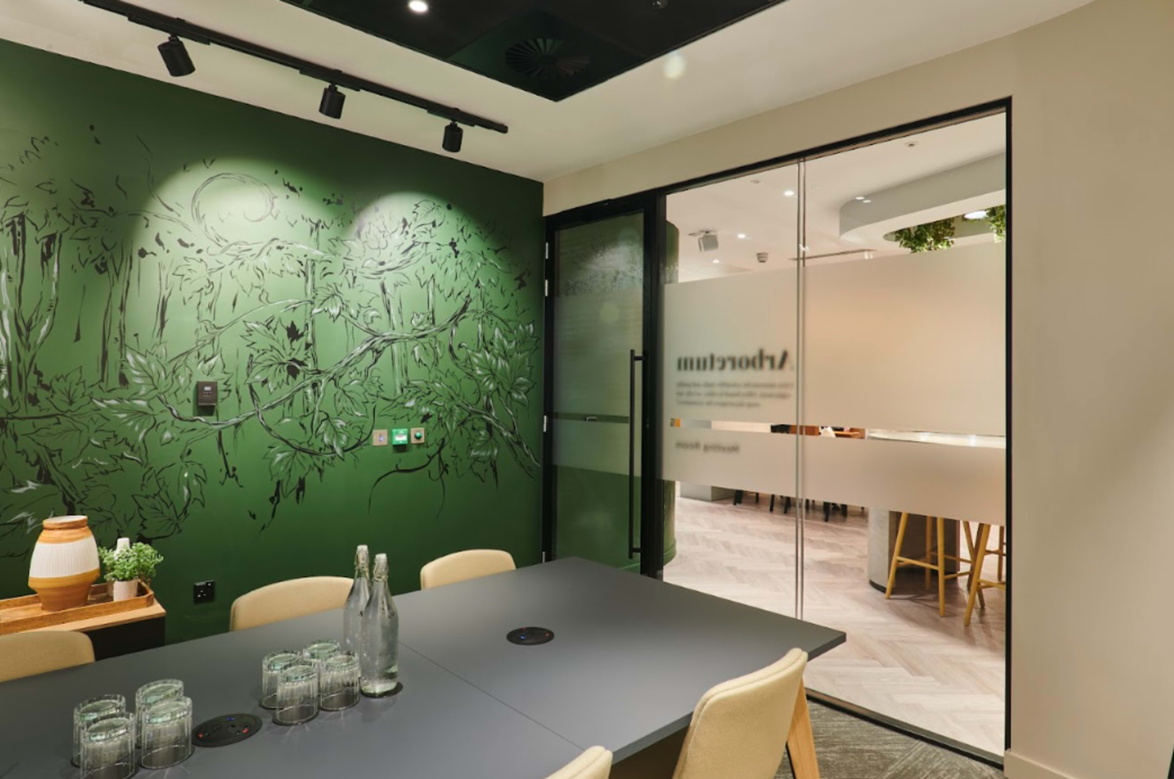 A modern meeting room with a dark green wall featuring botanical artwork and glass doors leading to an adjacent space.