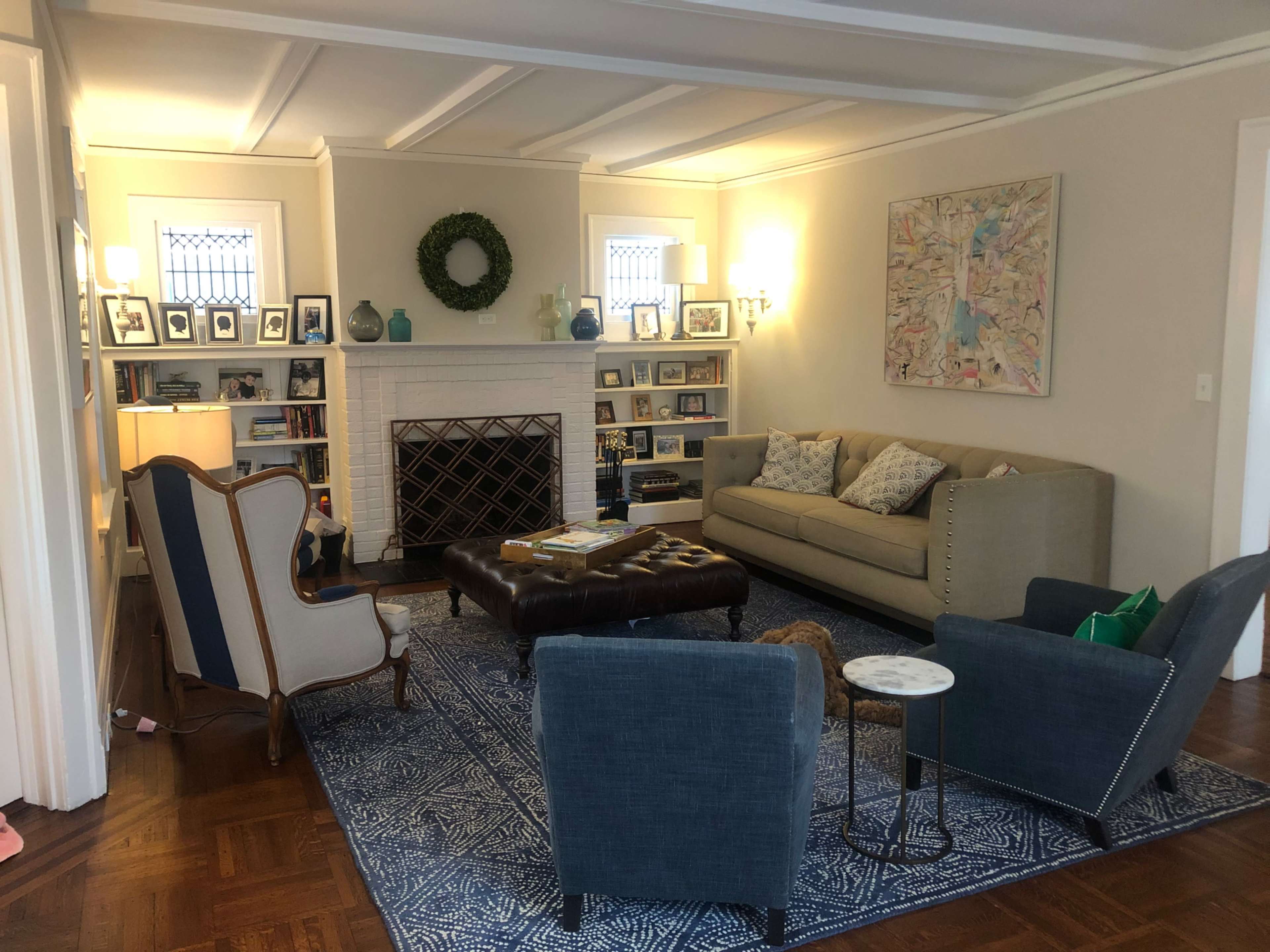 The image shows a living room with a fireplace, a sofa, two armchairs, a coffee table, and various framed photographs on display.