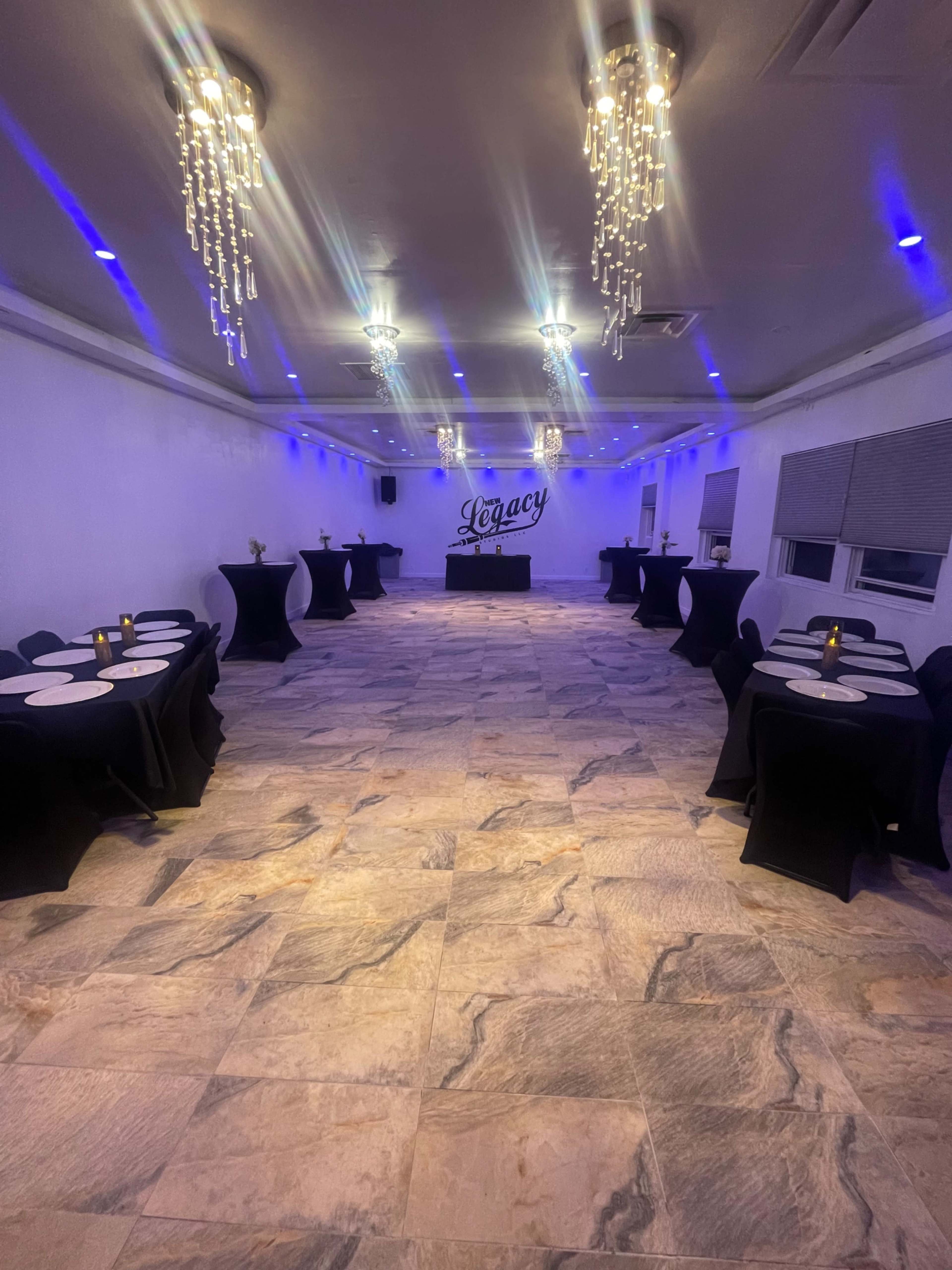 The room features a spacious layout with round tables covered in black cloth, illuminated by blue lights and adorned with hanging chandeliers.