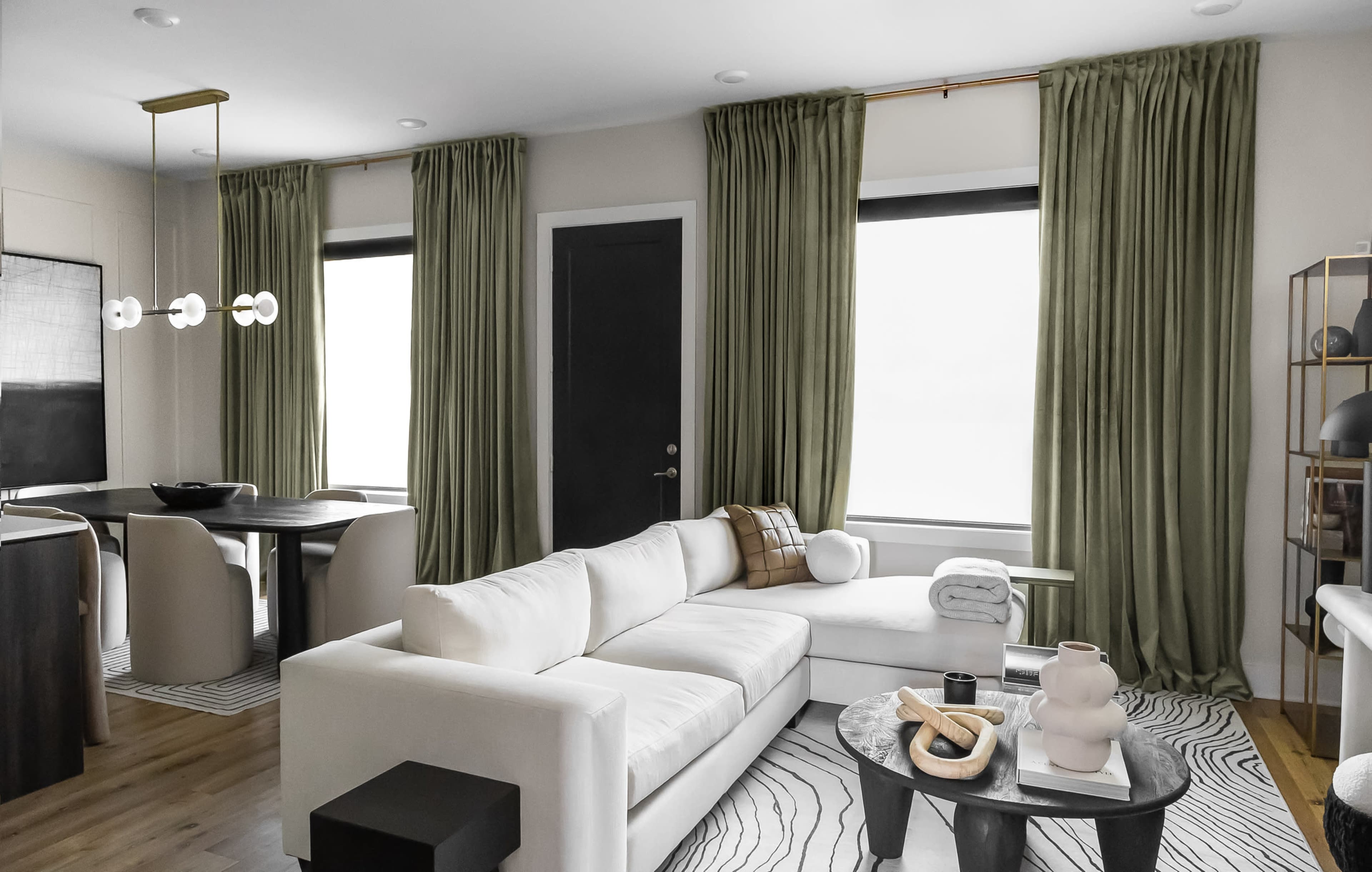 A modern living room features a white sectional sofa, a round black coffee table, and large windows dressed with olive-green curtains.