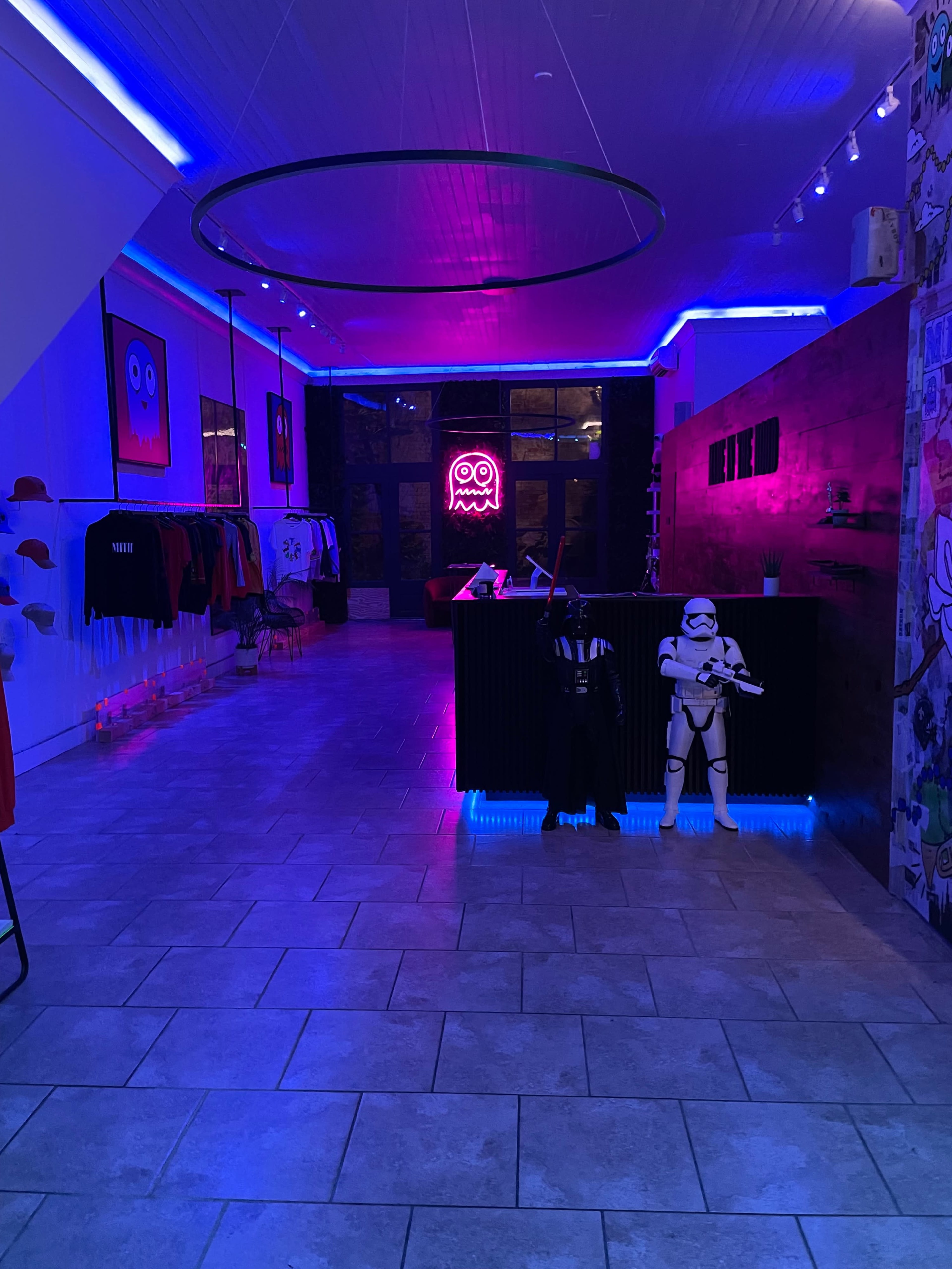 A modern retail space with colorful neon lighting, featuring clothing racks and a reception desk, alongside a stormtrooper figure.