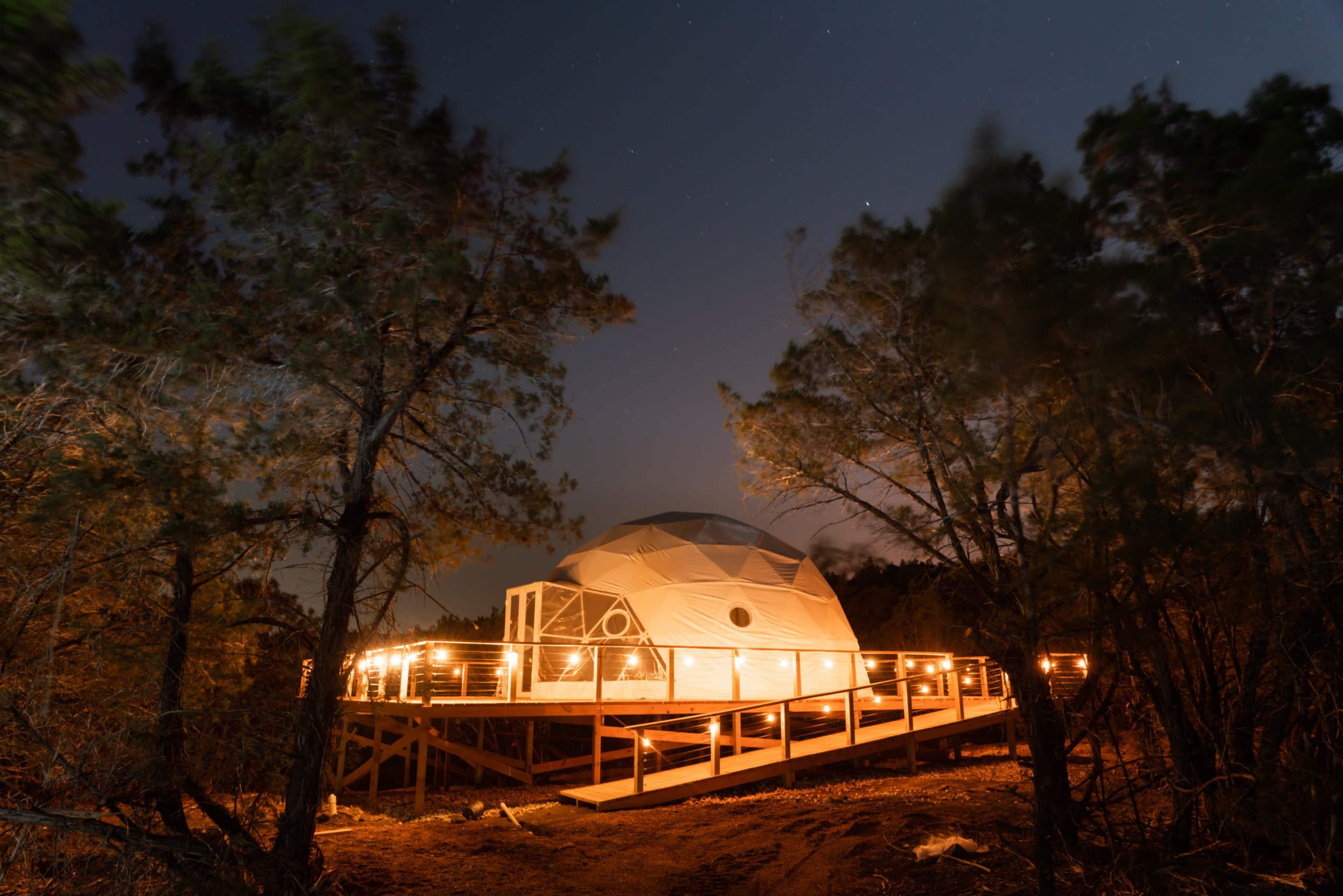 A geodesic dome on a raised platform is illuminated at night, surrounded by trees under a starry sky.