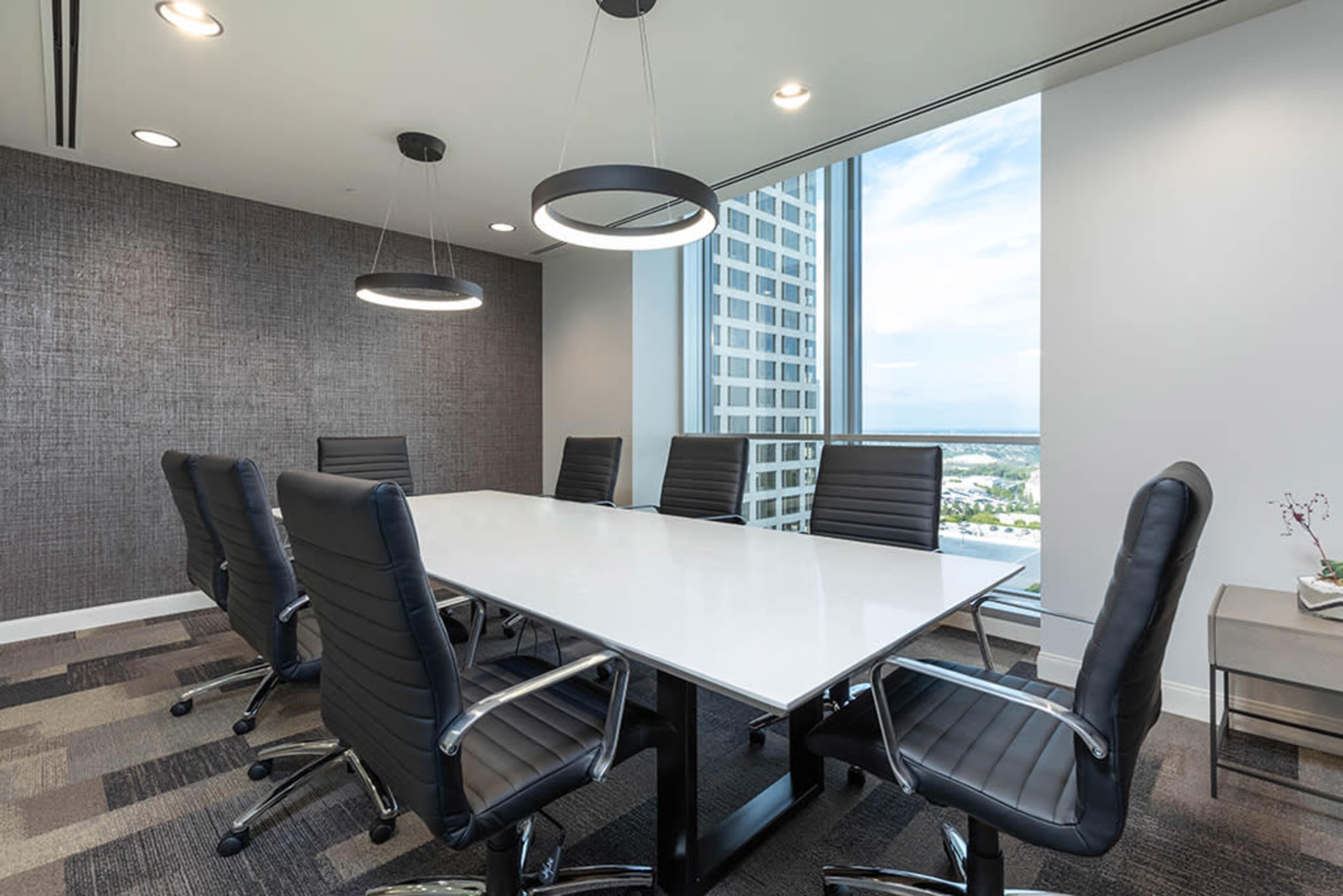 A modern conference room features a white rectangular table surrounded by eight black cushioned chairs, with large windows providing a view of the city skyline.
