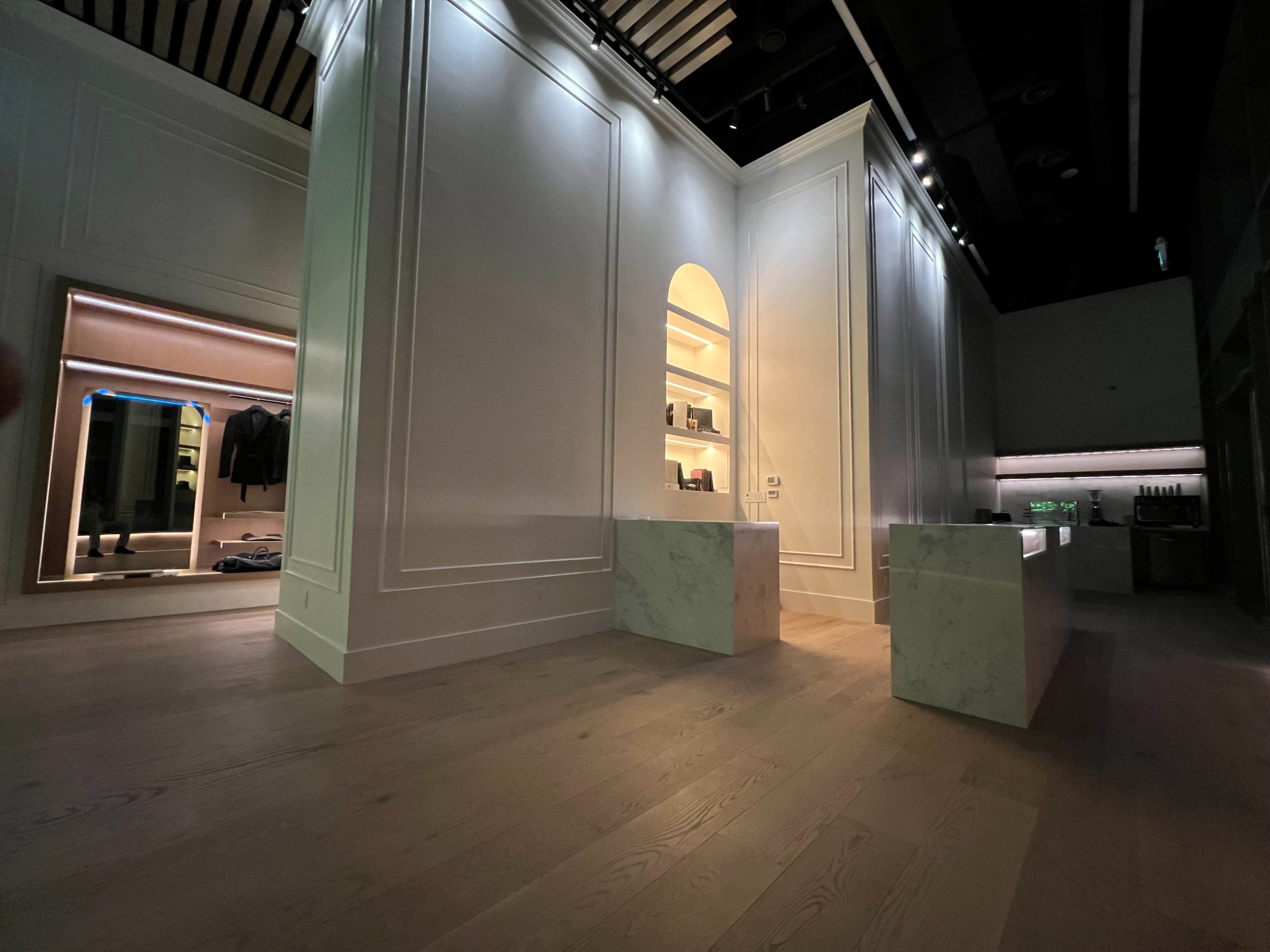 The image shows a minimalist interior space with white walls, marble counters, and a softly lit area displaying shelves, creating a modern, open layout.