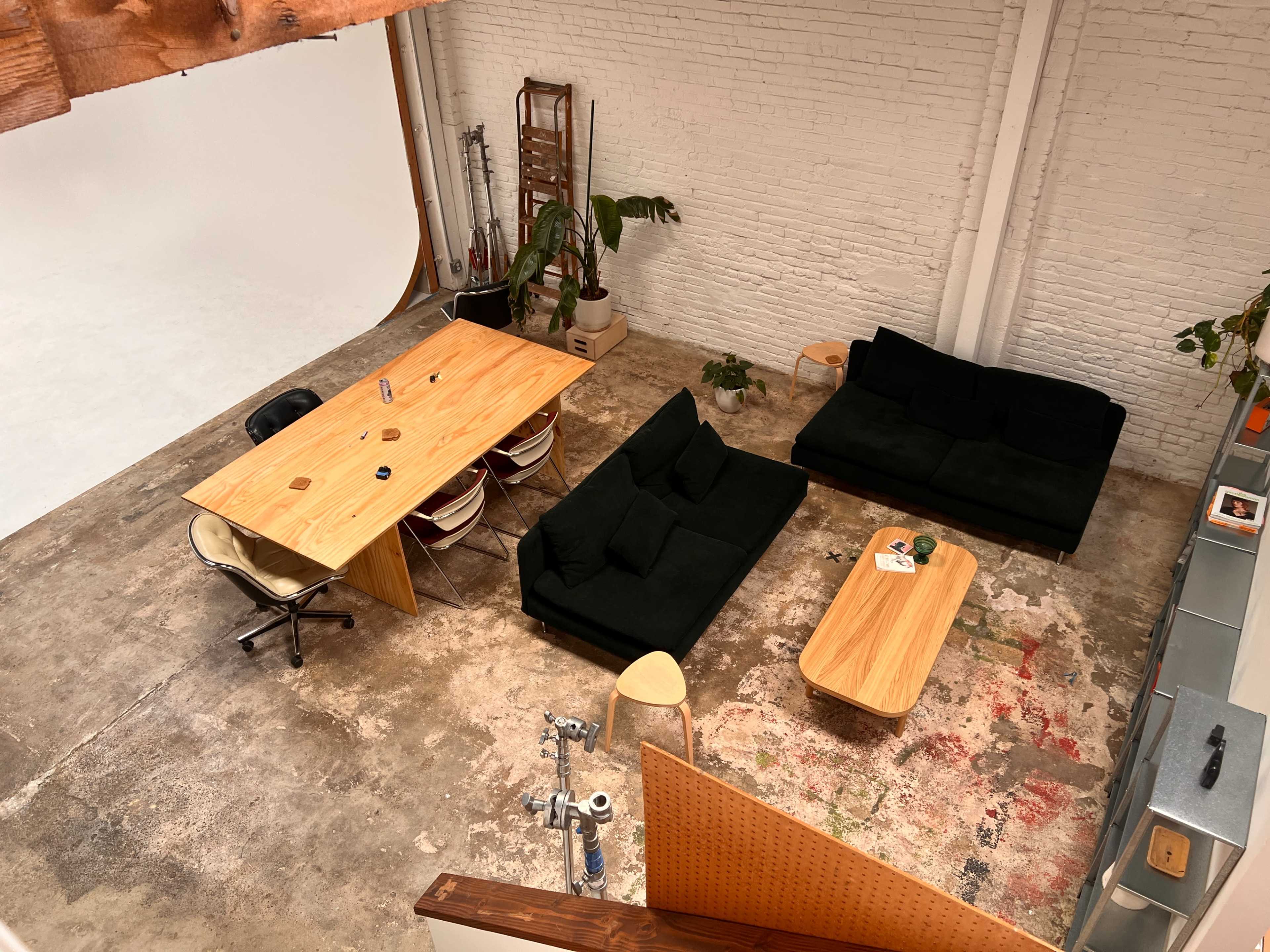 A spacious, industrial-style room with a wooden dining table surrounded by chairs, two dark sofas, a coffee table, and various plants, set against a backdrop of white brick walls.