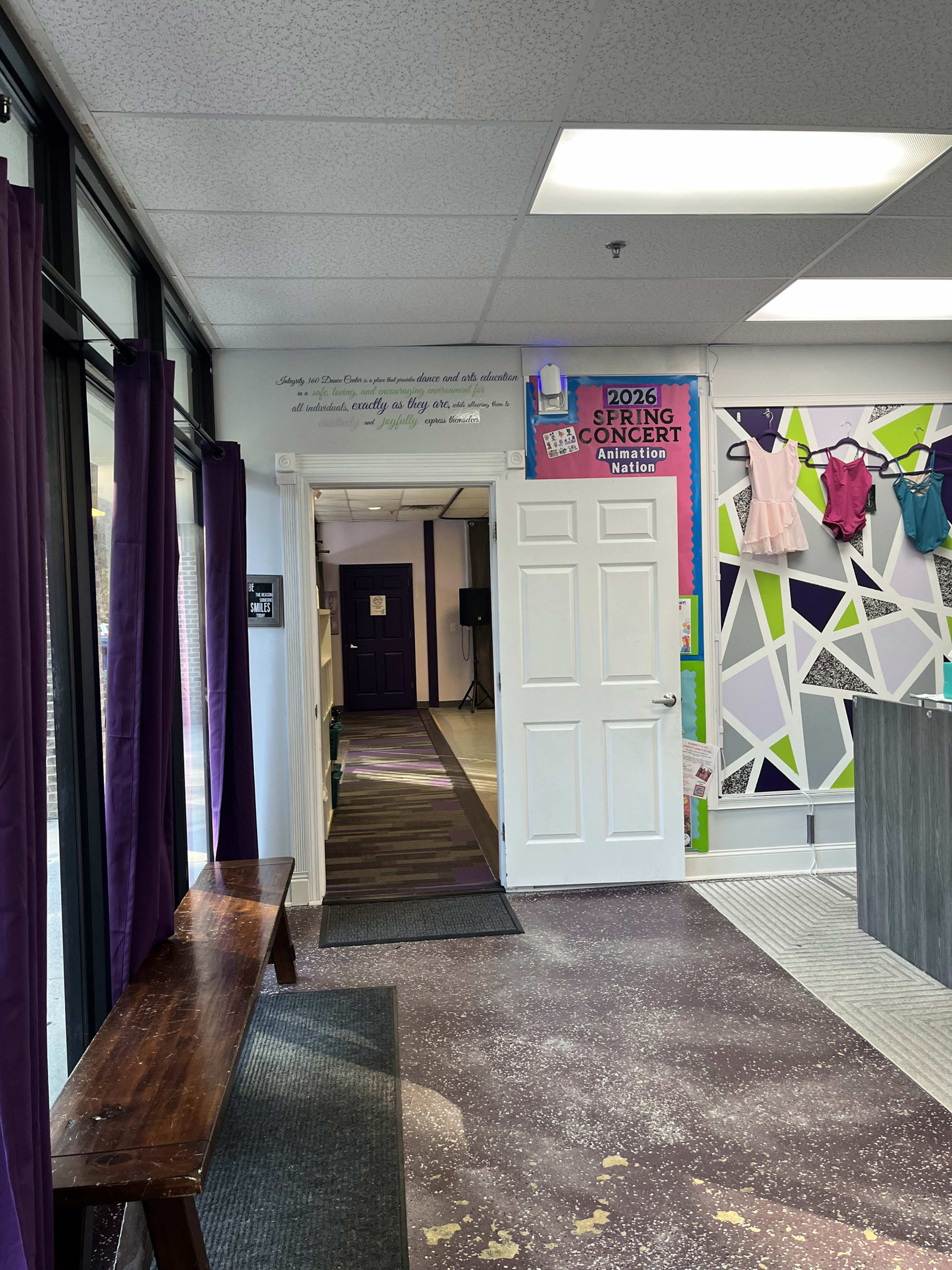 The image shows a hallway with a bench on the left, leading to a purple door at the end, and walls adorned with colorful posters and garments displayed on hangers.