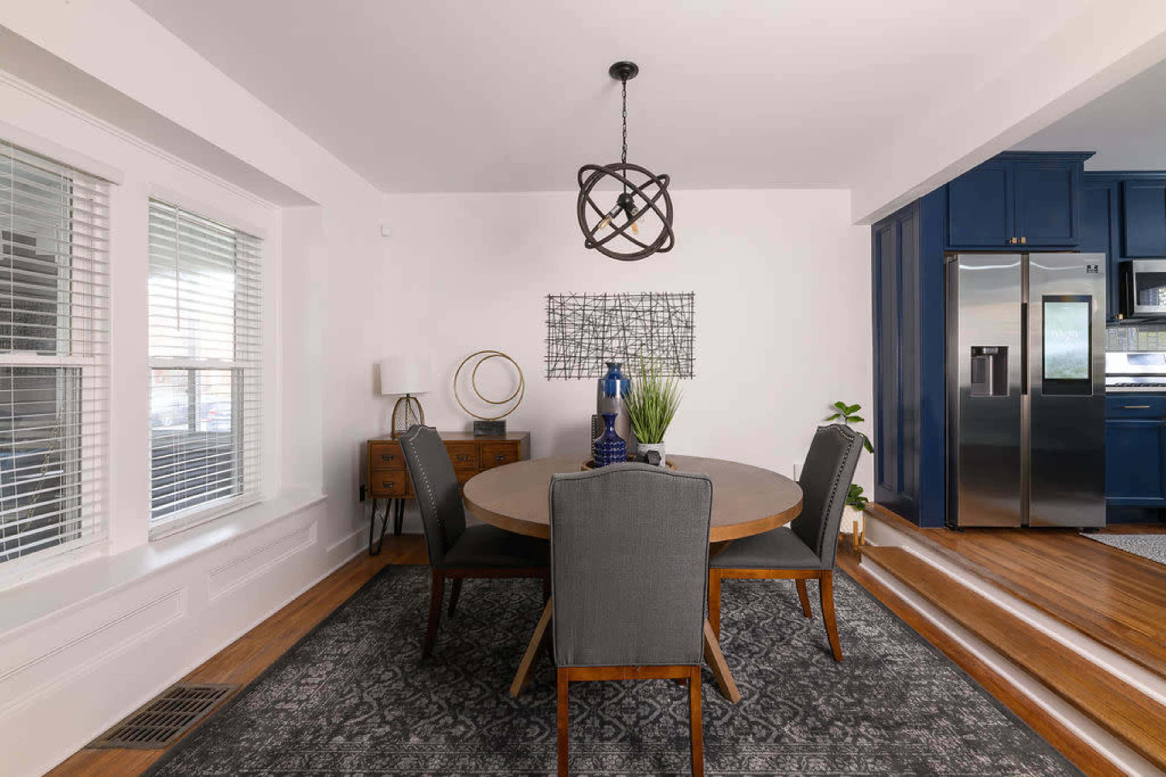 A round wooden dining table is set in a well-lit room with large windows, surrounded by four upholstered chairs, a decorative pendant light hanging above, and a kitchen area visible in the background.