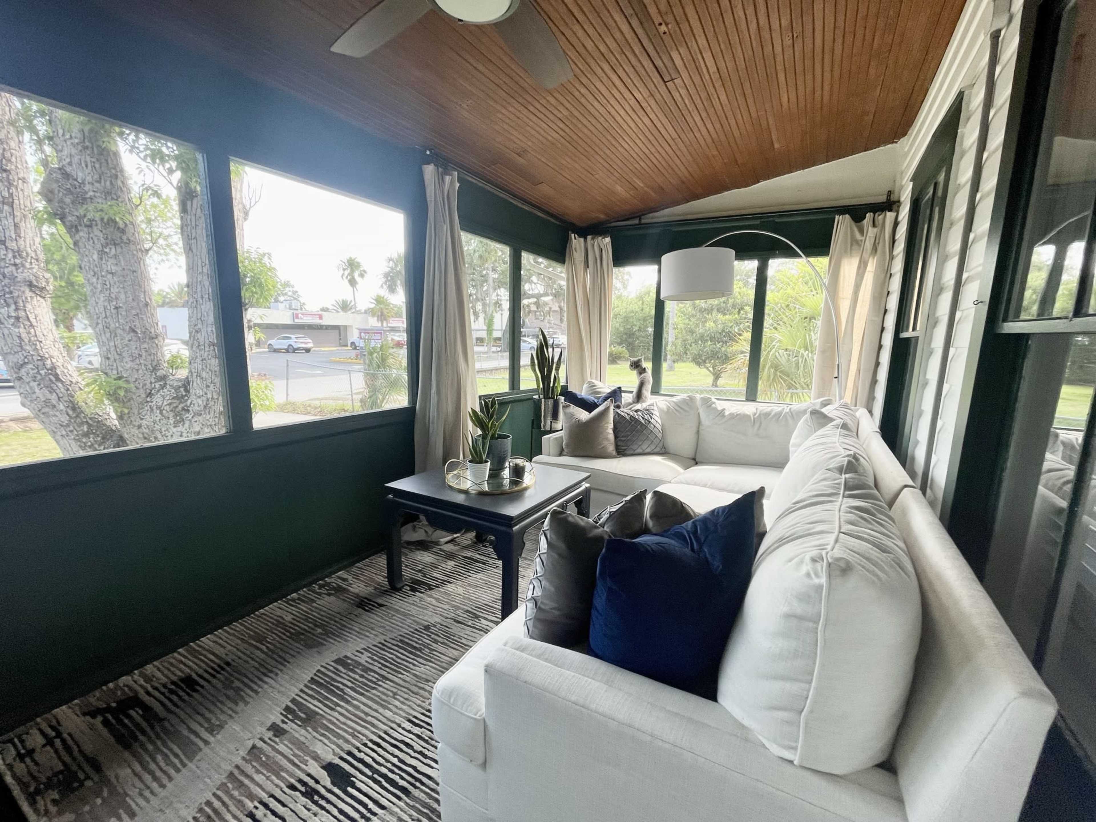 A screened porch features a white sectional sofa and a dark coffee table, with large windows framing a view of greenery outside.