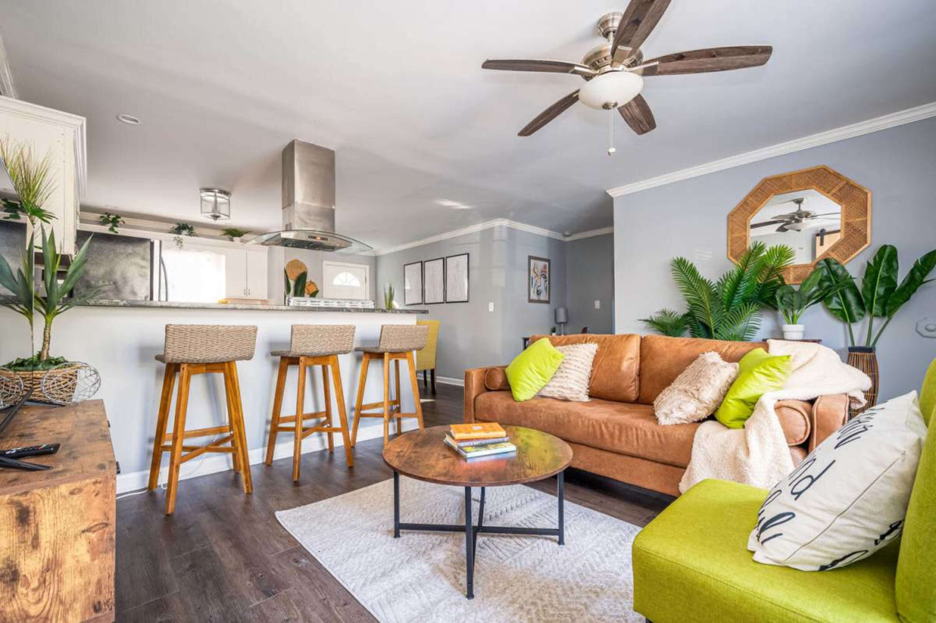 The image shows a modern living room with a brown sofa, a round coffee table, and a kitchen area in the background, featuring tall bar stools and a stainless steel range hood.