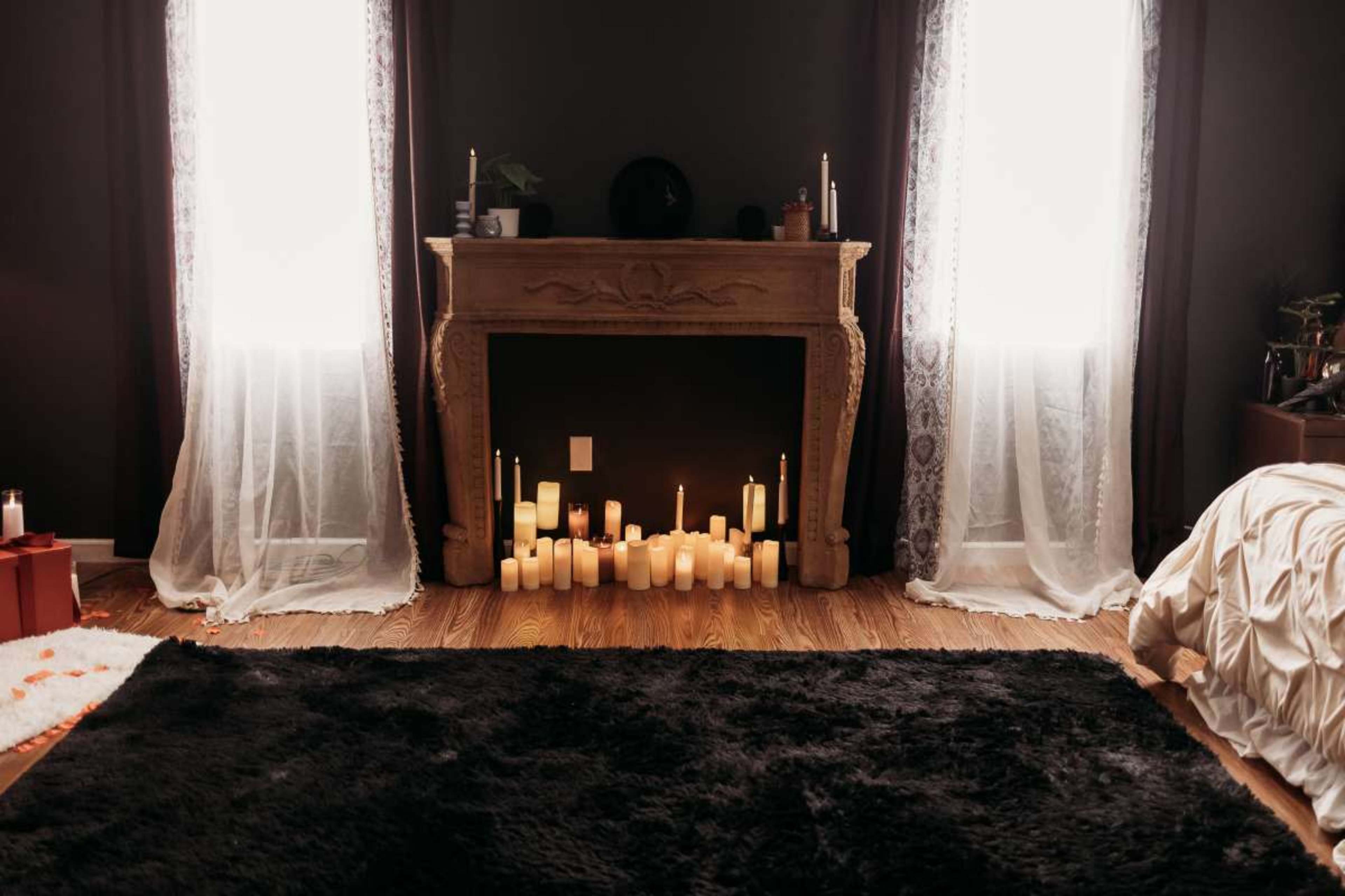 The room features a dark color scheme with a decorative fireplace surrounded by numerous candles, flanked by two windows draped with sheer curtains.